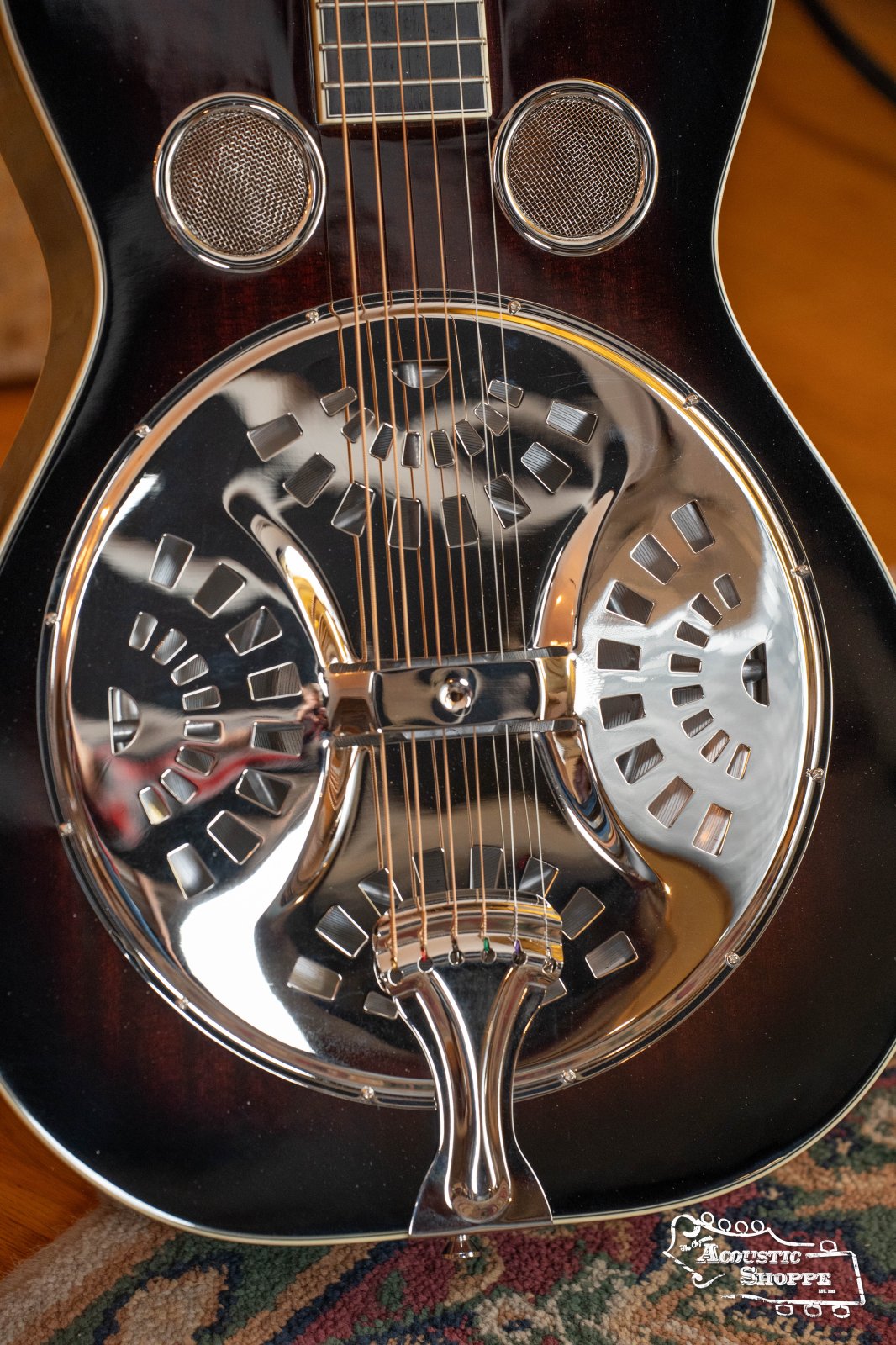Close-up of the Recording King RR-36S-VS Maxwell Series #4706 resonator guitar showing its shiny metal cover, decorative sound holes, hand-spun European cone, strings, and fretboard. The guitar rests on a surface with a patterned rug below.