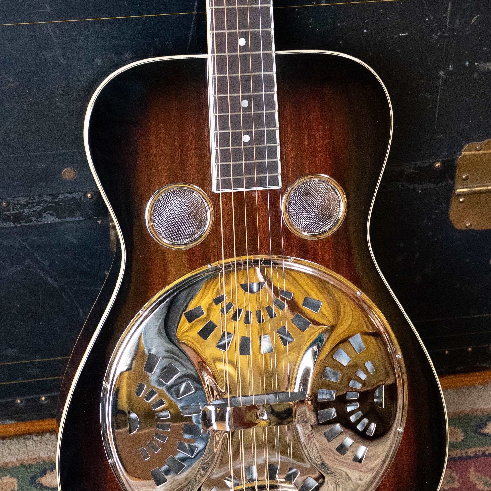A Recording King RR-36S-VS All Mahogany Vintage Sunburst Square Neck Resonator guitar with a shiny metal resonator plate and two covered sound holes leans against a dark vintage trunk.