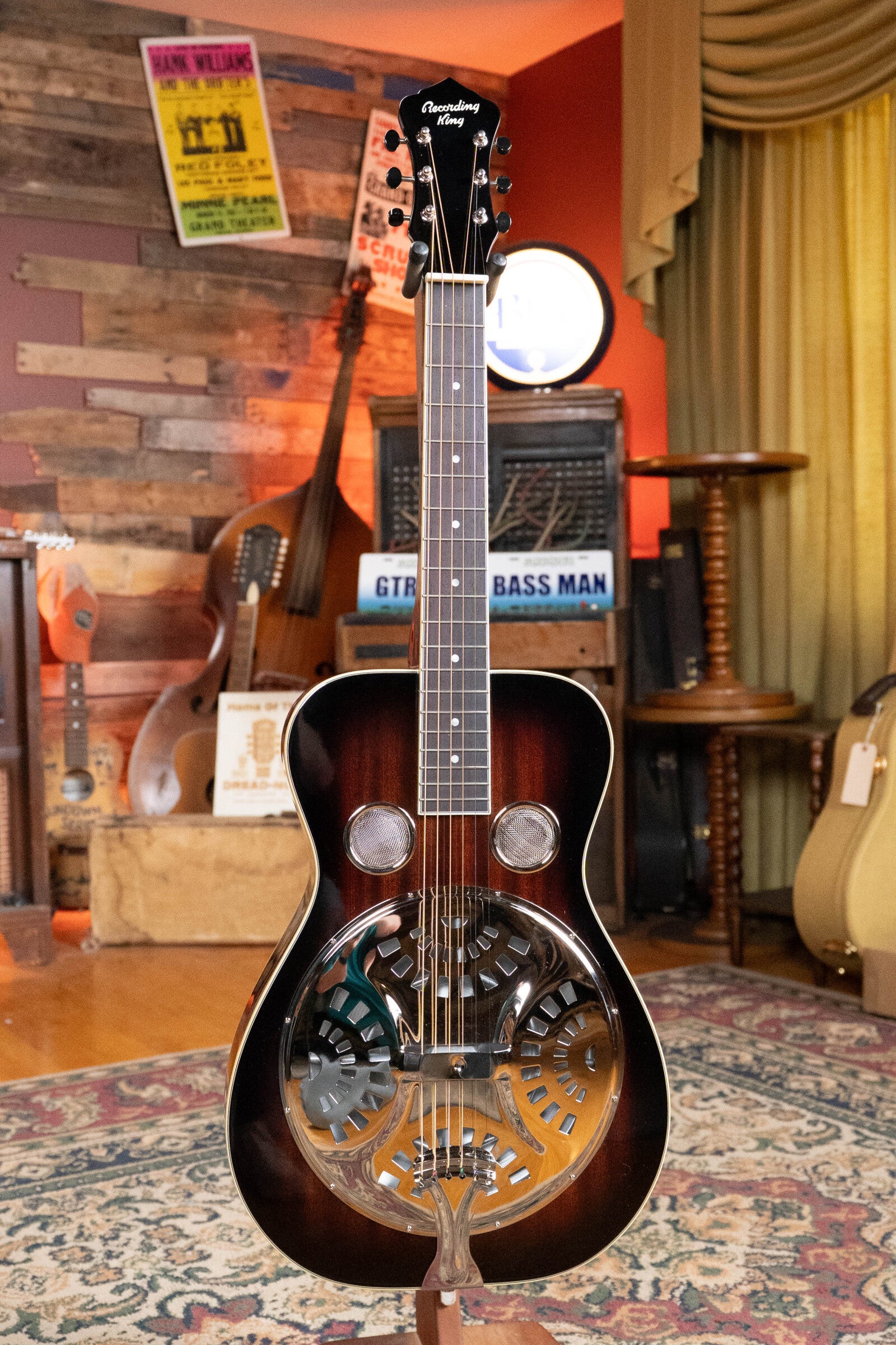 The Recording King RR-36S-VS All Mahogany Vintage Sunburst Square Neck Resonator from the Maxwell Series (#6003) stands upright in a cozy music room, among other string instruments, amps, and music posters on wooden walls.