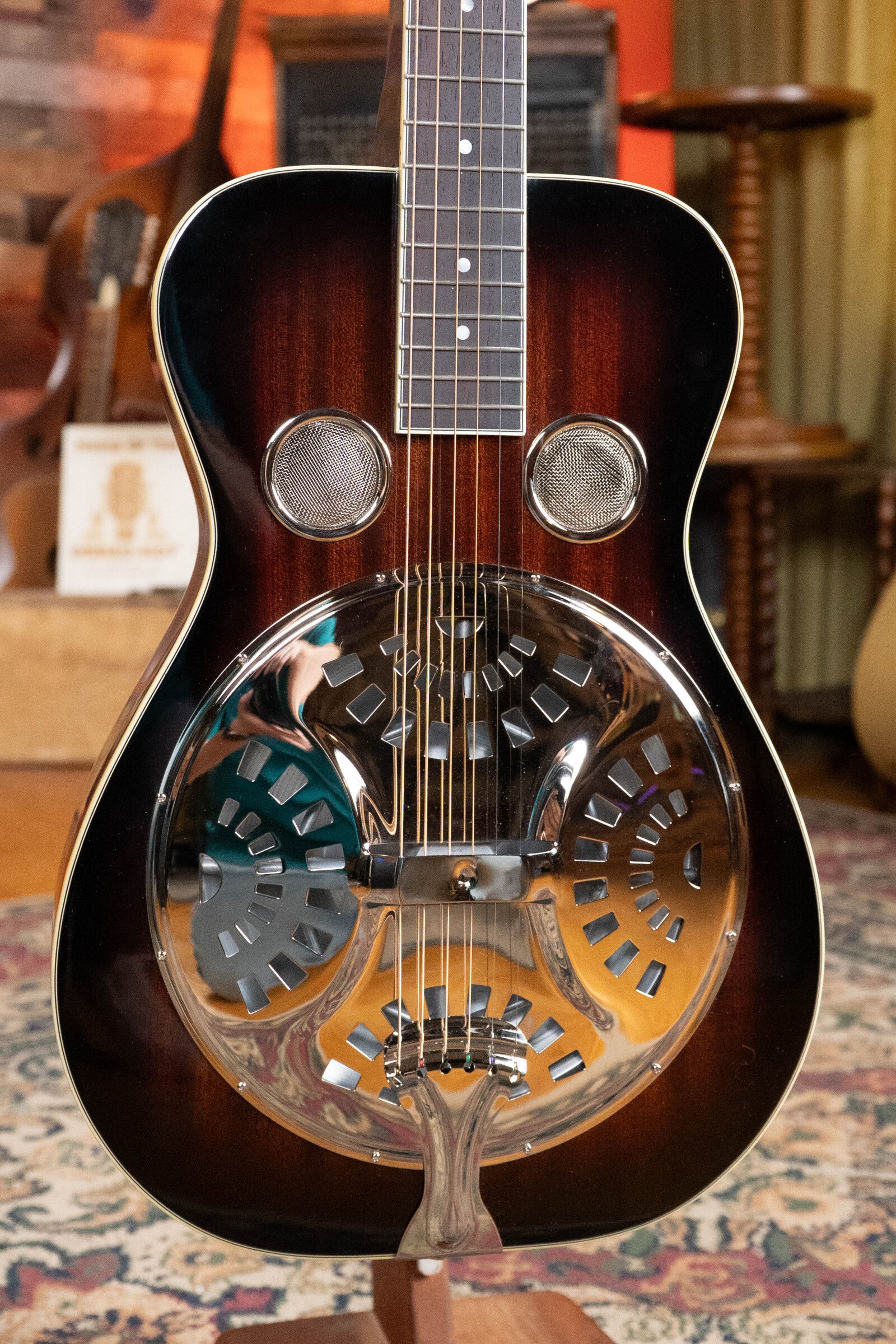 A close-up of the Recording King RR-36S-VS All Mahogany Vintage Sunburst Square Neck Resonator Maxwell Series #6003, featuring a mahogany body, two circular sound holes, and dark sunburst finish, displayed indoors on a patterned rug.