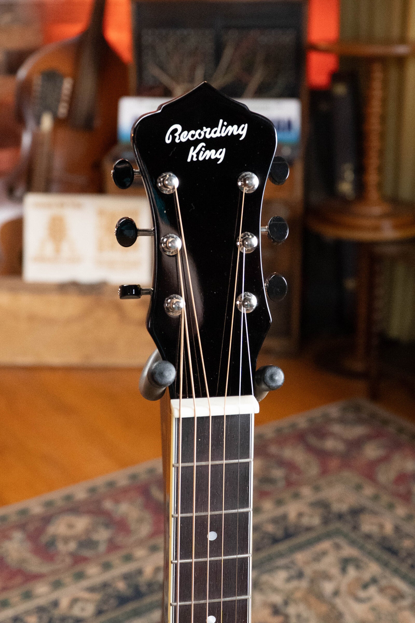 Close-up of the headstock of a Recording King RR-36S-VS All Mahogany Vintage Sunburst Square Neck Resonator Maxwell Series #6003, showing tuning pegs and logo with a blurred background of books, furniture, and a patterned rug.