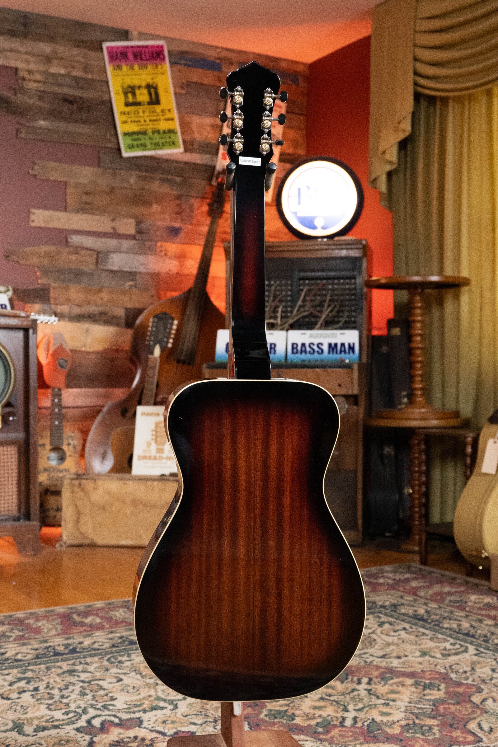 The Recording King RR-36S-VS All Mahogany Vintage Sunburst Square Neck Resonator Maxwell Series #6003 stands upright, back facing out, in a cozy room with vintage music posters, instruments, and a patterned rug under warm light.