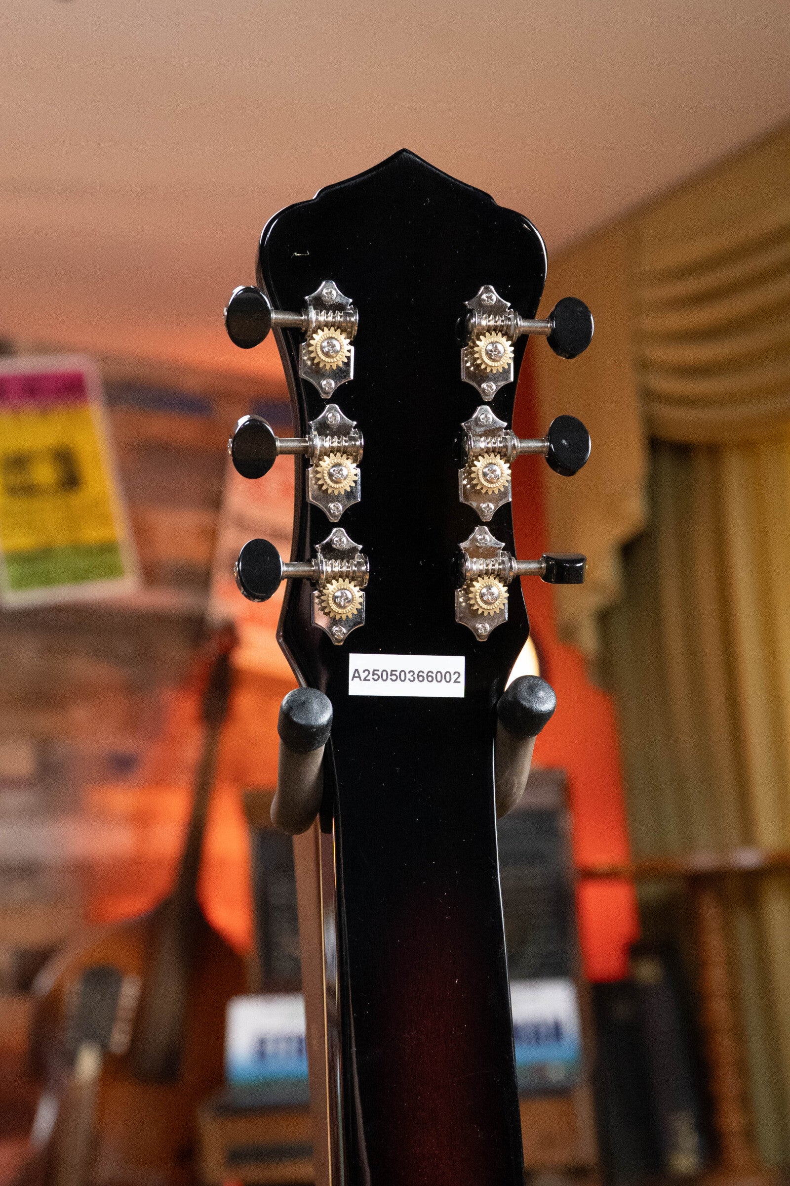 Close-up of the back of a Recording King RR-36S-VS All Mahogany Vintage Sunburst Square Neck Resonator’s headstock, showing six metal tuning pegs and a white barcode label; blurred instruments and posters fill the warmly lit room.