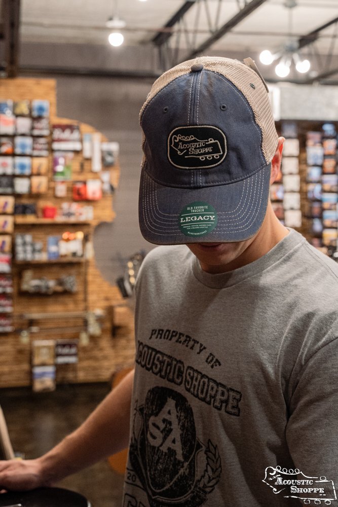 A person wearing an Amplify Screen Printing & Promotions Acoustic Shoppe Legacy Badge Trucker Cap in Royal Blue/Black stands indoors, head lowered, with musical accessories displayed on wooden shelves in the background.