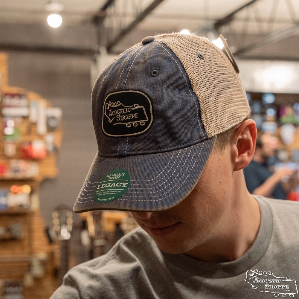 A person wearing an Amplify Screen Printing & Promotions Acoustic Shoppe Legacy Badge Trucker Cap in Royal Blue/Black stands indoors with head bowed, blurred shelves and acoustic guitars visible in the background.