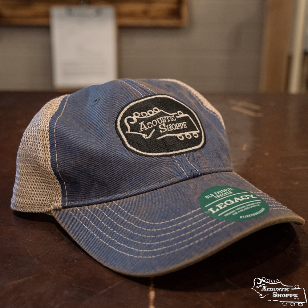 The Acoustic Shoppe Legacy Badge Trucker Cap by Amplify Screen Printing & Promotions features royal blue and black mesh with an embroidered front logo and a Legacy sticker on the brim, displayed on a dark wooden surface.