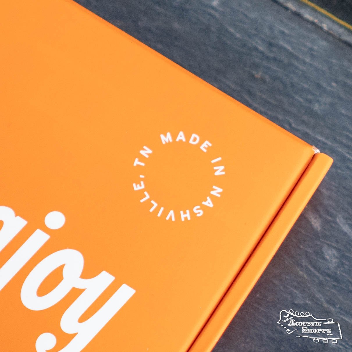 Close-up of an orange Stringjoy box with enjoy in white and a MADE IN NASHVILLE, TN stamp. The box holds Stringjoy Mandolin Foxwoods Medium Gauge (11-40) Coated Phosphor Bronze Strings. Part of a gray surface appears in the background.