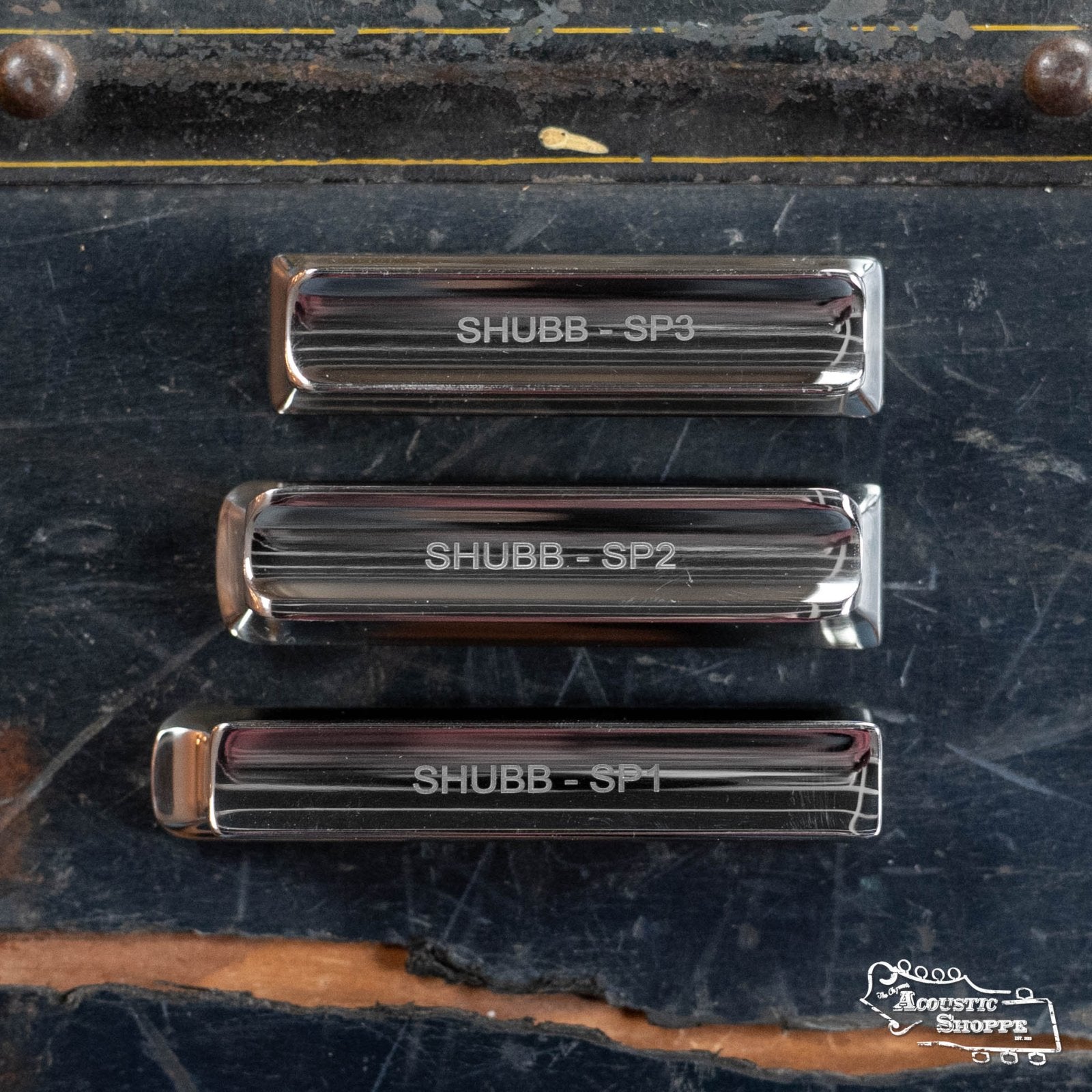 The Shubb Pearse Slide (SP2) by Shubb is displayed vertically on a worn dark surface with The Acoustic Shoppe logo, ideal for resophonic or lap steel guitar players seeking a high-quality steel slide.