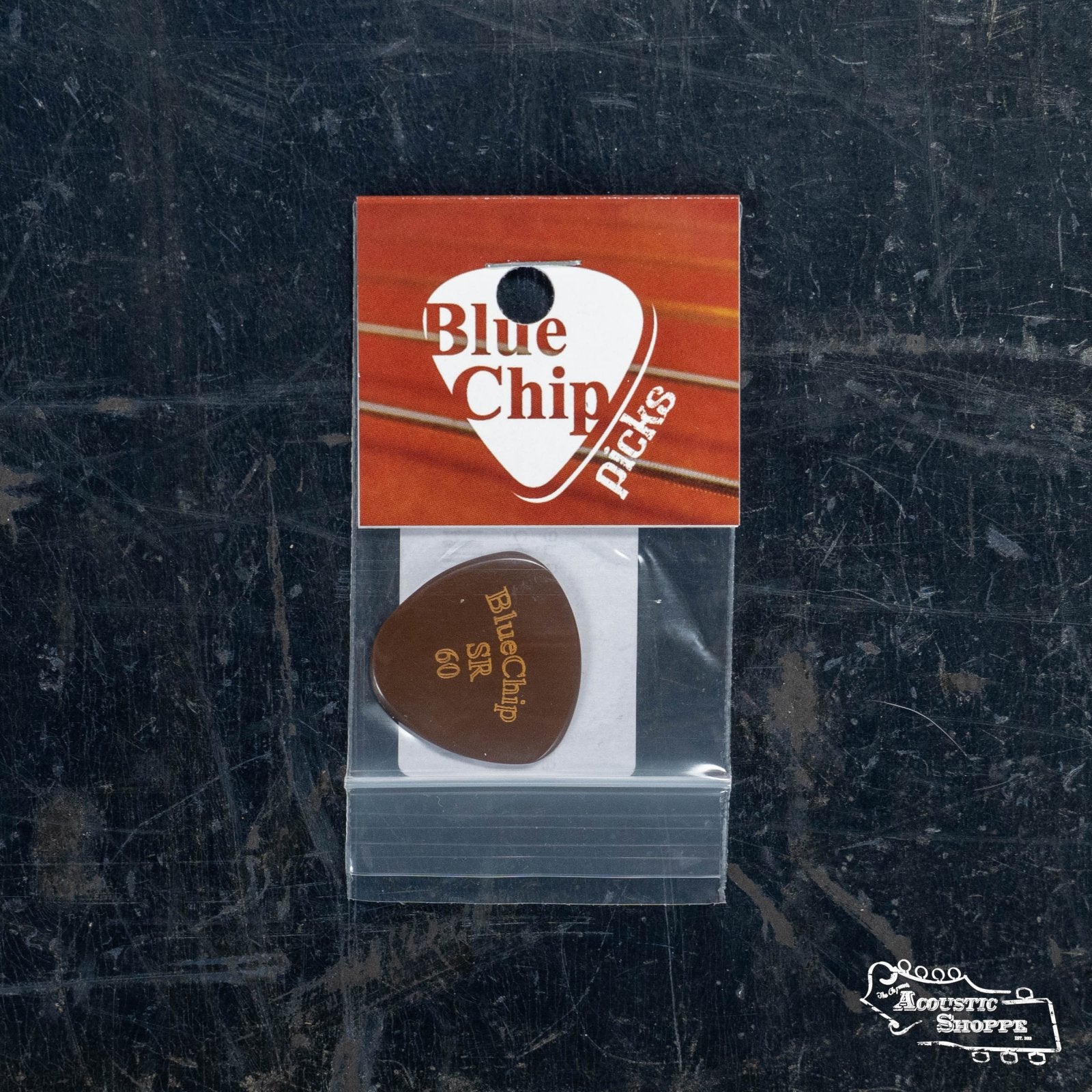 A BlueChip Picks (SR60) guitar pick by BlueChip is packaged in a clear plastic bag on an orange and white card labeled “BlueChip Picks,” featuring the Acoustic Shoppe logo, and rests on a dark scratched surface.