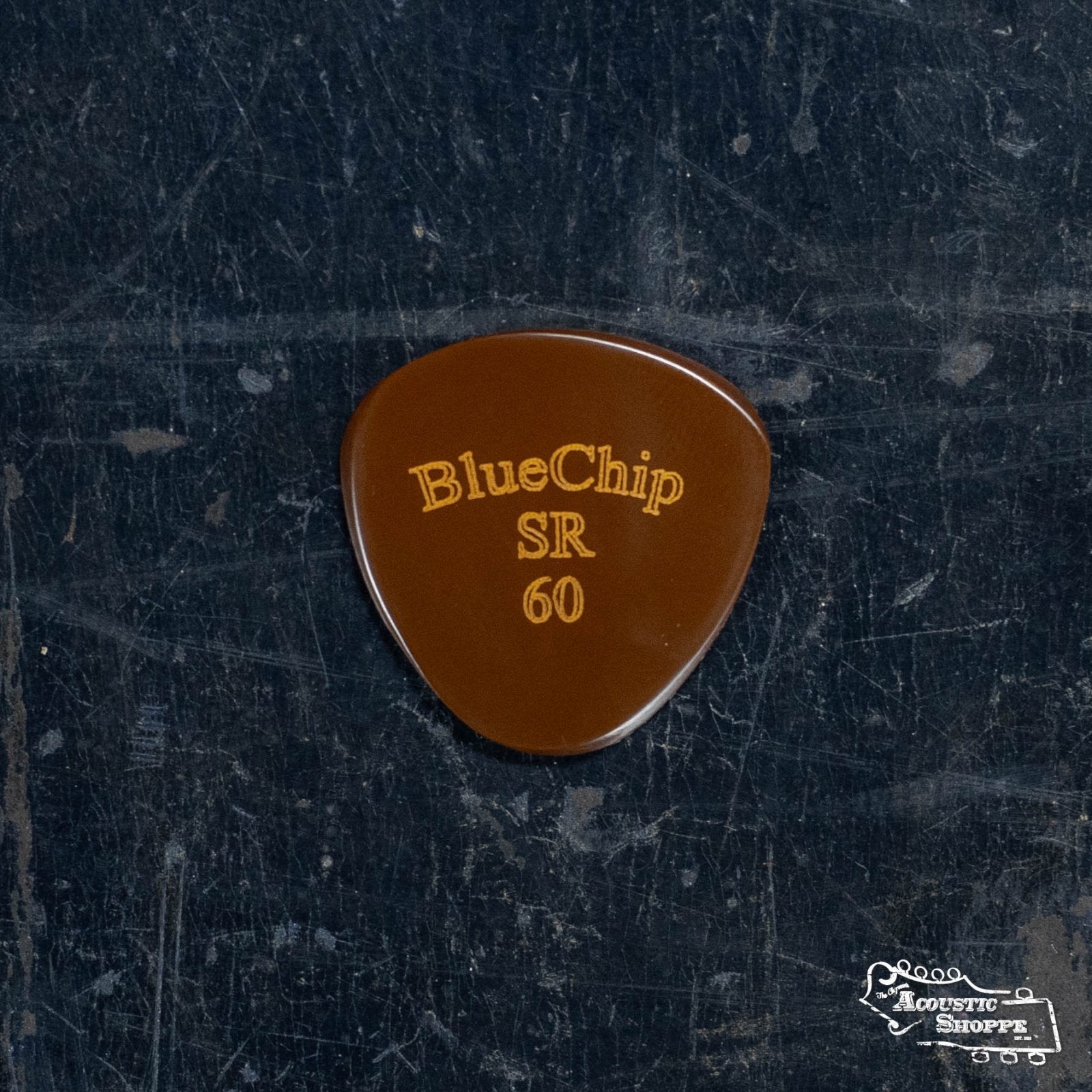 A brown, precision-machined BlueChip Picks (SR60) guitar pick with gold lettering rests on a dark, textured surface. The Acoustic Shoppe logo appears in the corner—a standout BlueChip accessory for acoustic musicians.