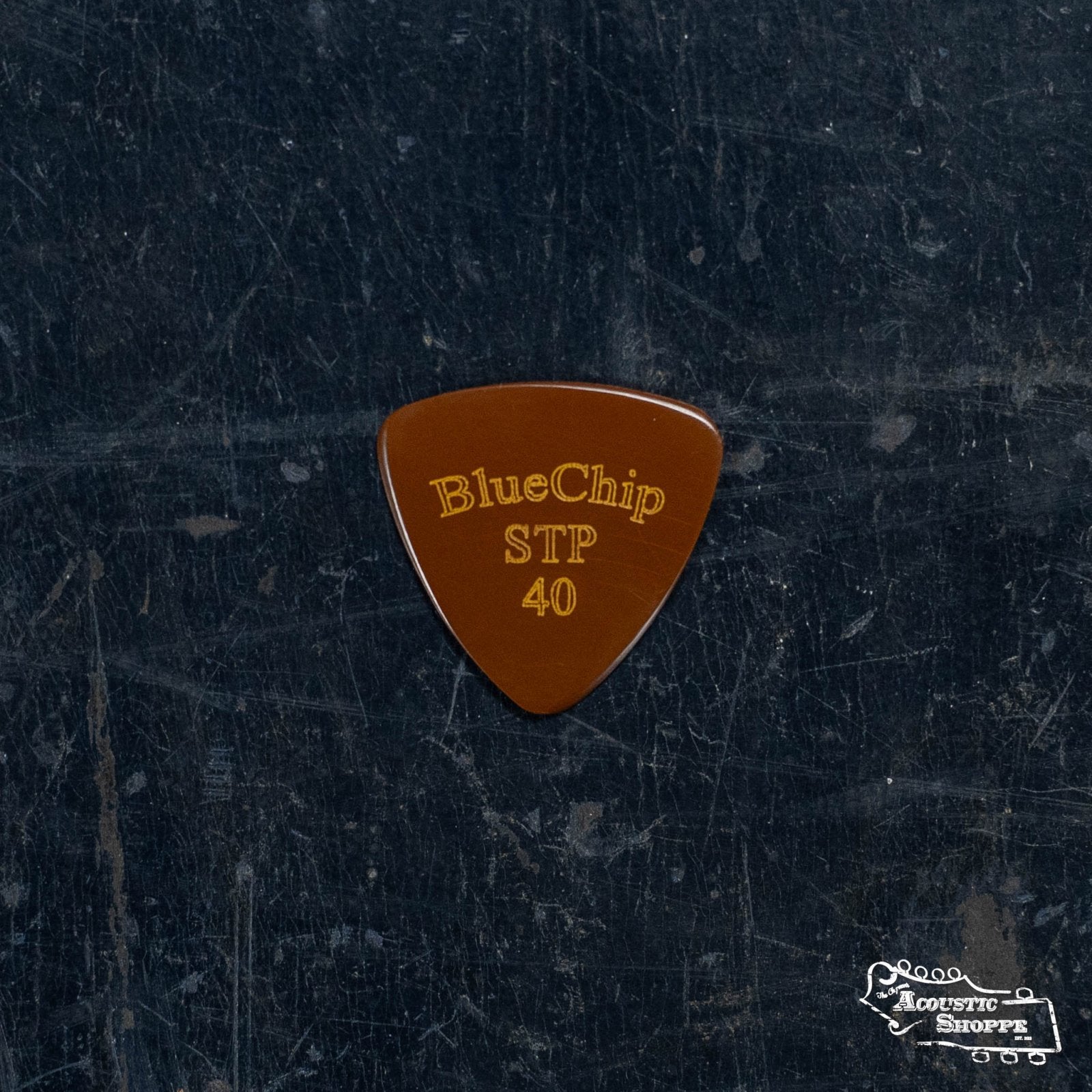 A brown BlueChip Picks (STP40) guitar pick with yellow STP 40 engraving rests on a dark textured surface. The Acoustic Shoppe logo appears at the lower right, highlighting this premium BlueChip accessory for acoustic musicians.