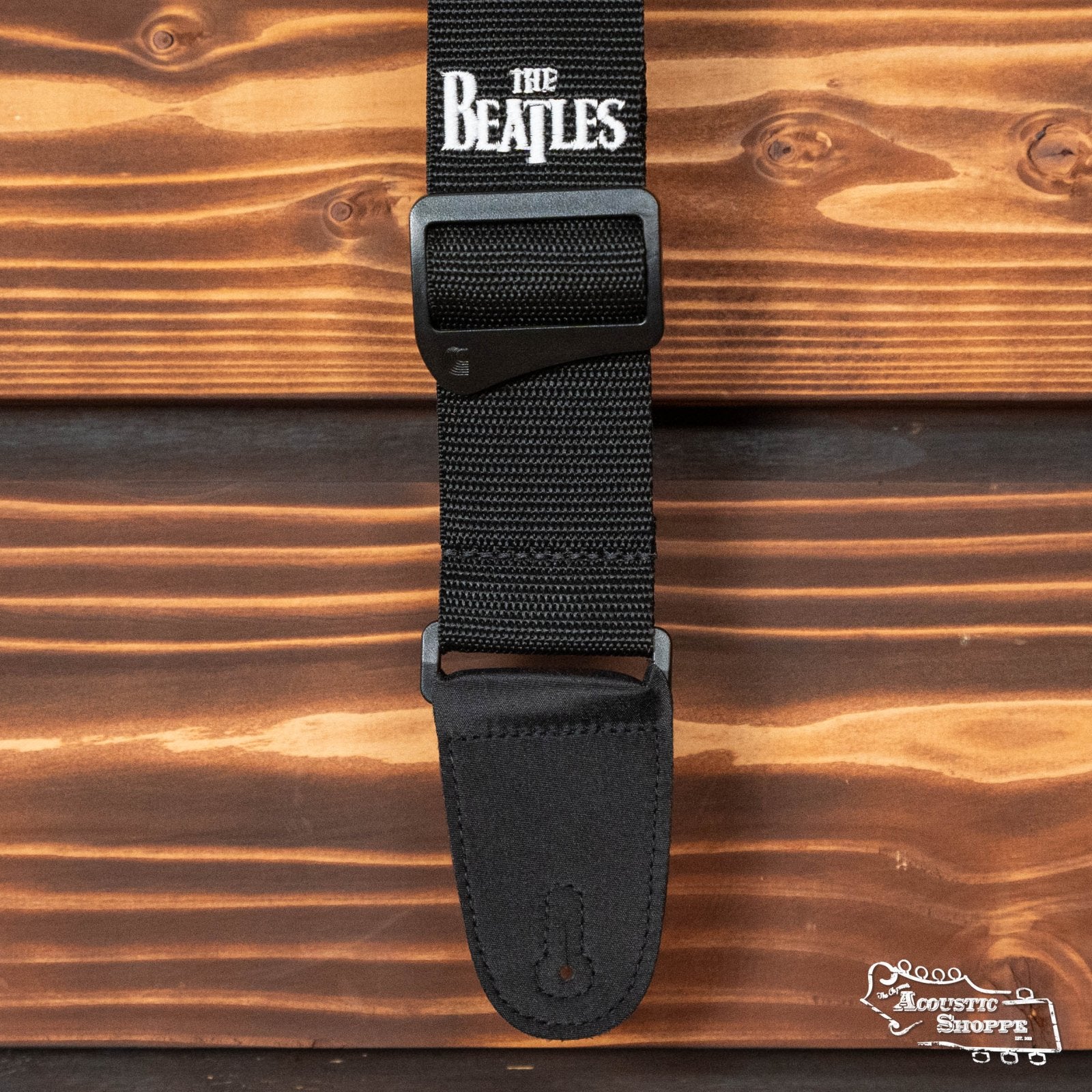The DAddario Beatles Polypropylene Guitar Strap in black, featuring The Beatles logo in white, is displayed against a wooden background. The Acoustic Shoppe logo appears in the bottom right corner.