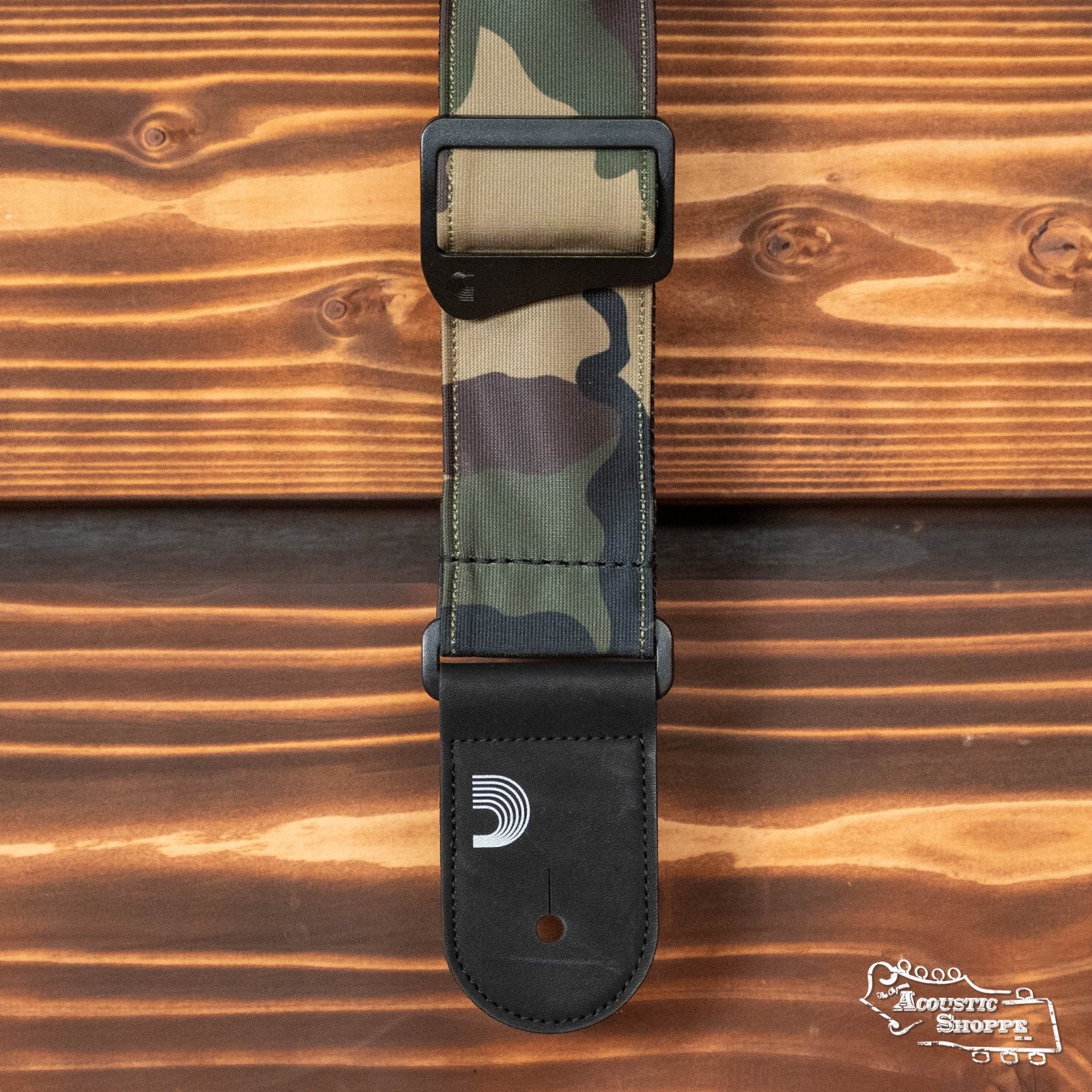 The DAddario Camouflage Guitar Strap, featuring black leather ends and a white logo, is displayed on a wooden surface with horizontal grain patterns.
