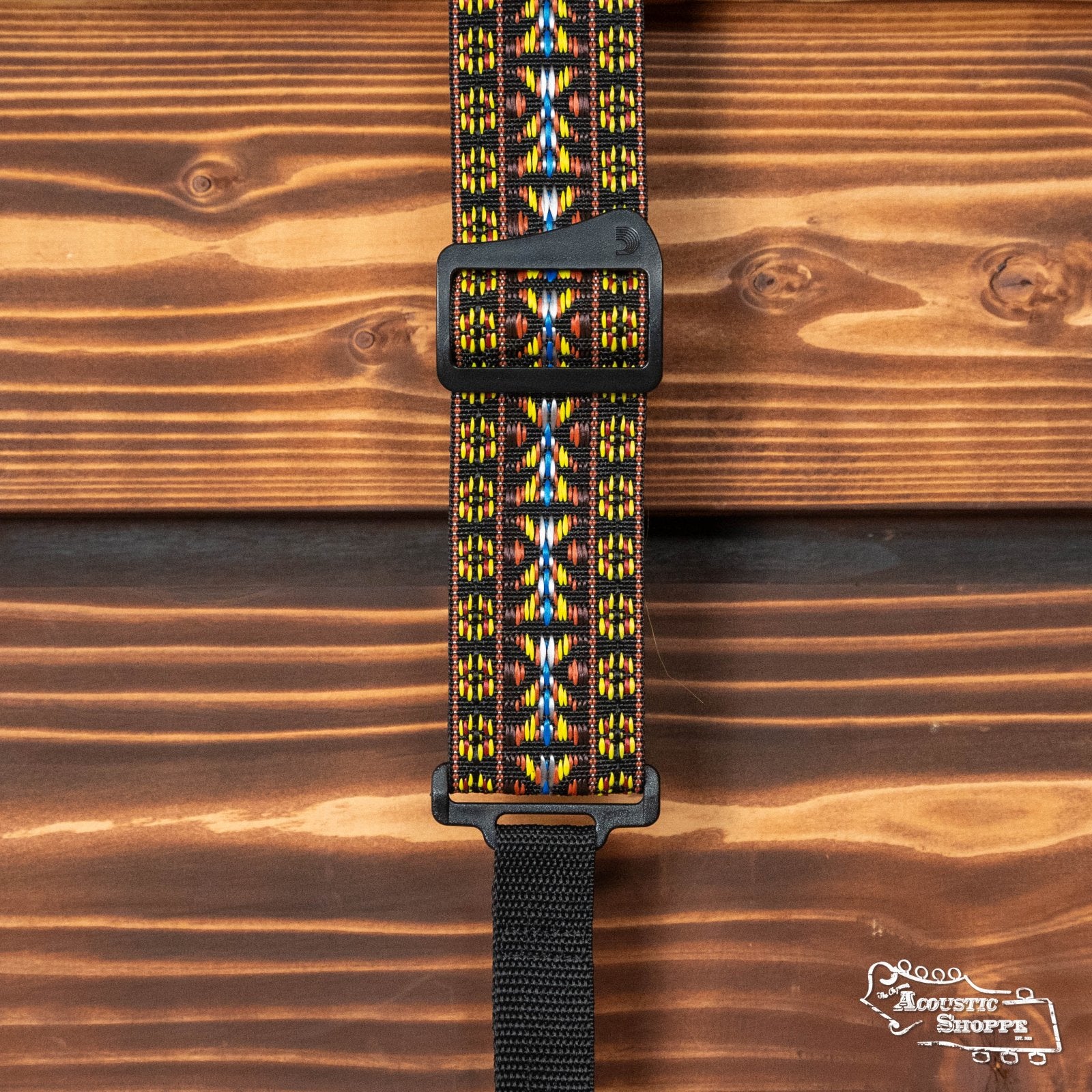 The DAddario 20N01BJ Woven Hootenanny Banjo Strap in yellow and brown features a geometric design, adjustable black plastic buckle, and is shown on a wooden background.