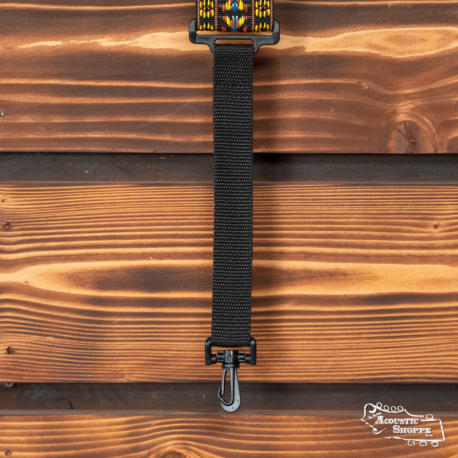 The DAddario 20N01BJ Woven Hootenanny Banjo Strap in yellow and brown features a colorful woven design and metal hook, hanging against a wooden wall. The Acoustic Shoppe logo appears in the bottom right corner.