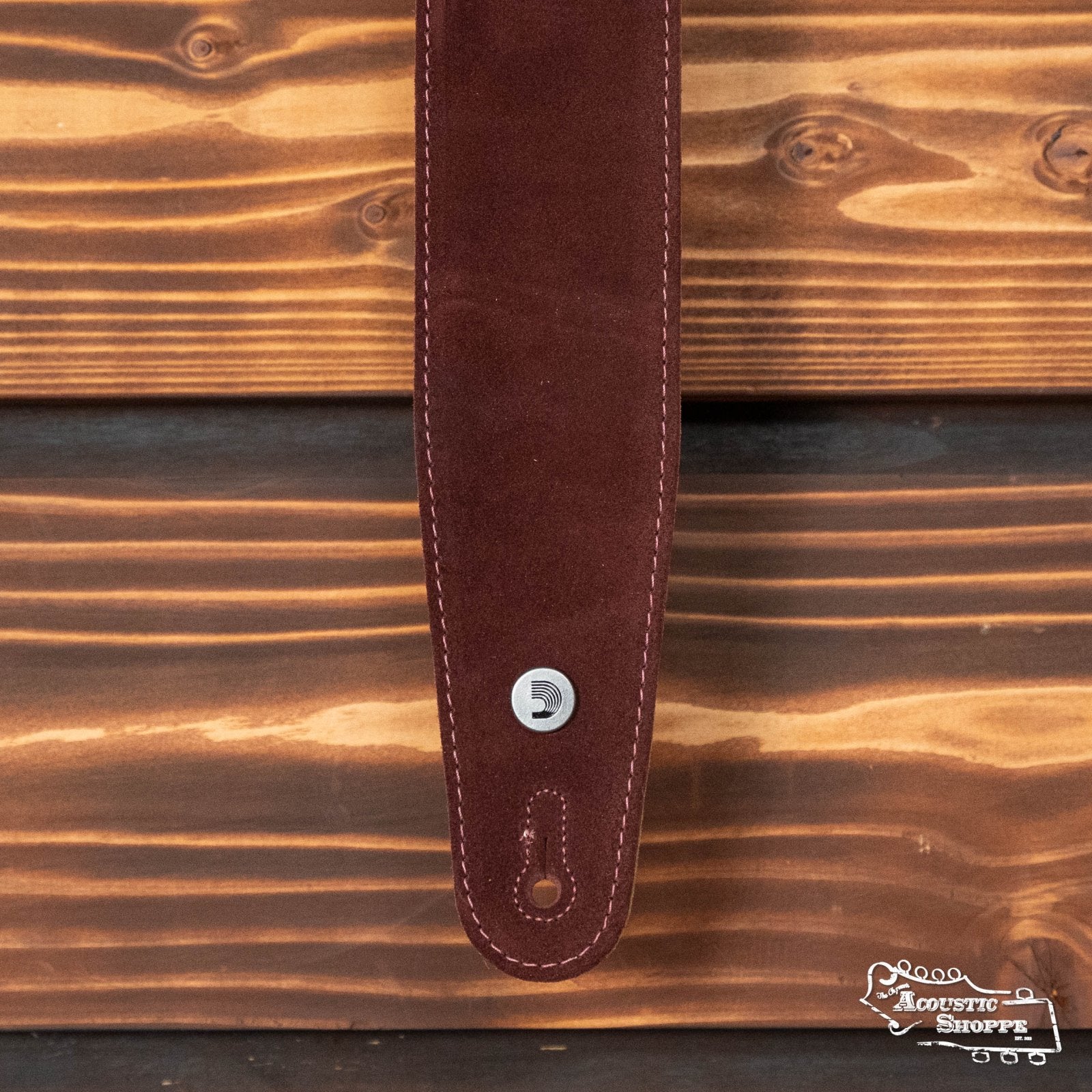 A close-up of the D'Addario Suede Guitar Strap in burgundy (25SS03-DX) with white stitching, metal button, and attachment hole on a wooden background. The Acoustic Shoppe logo appears in the lower right corner.