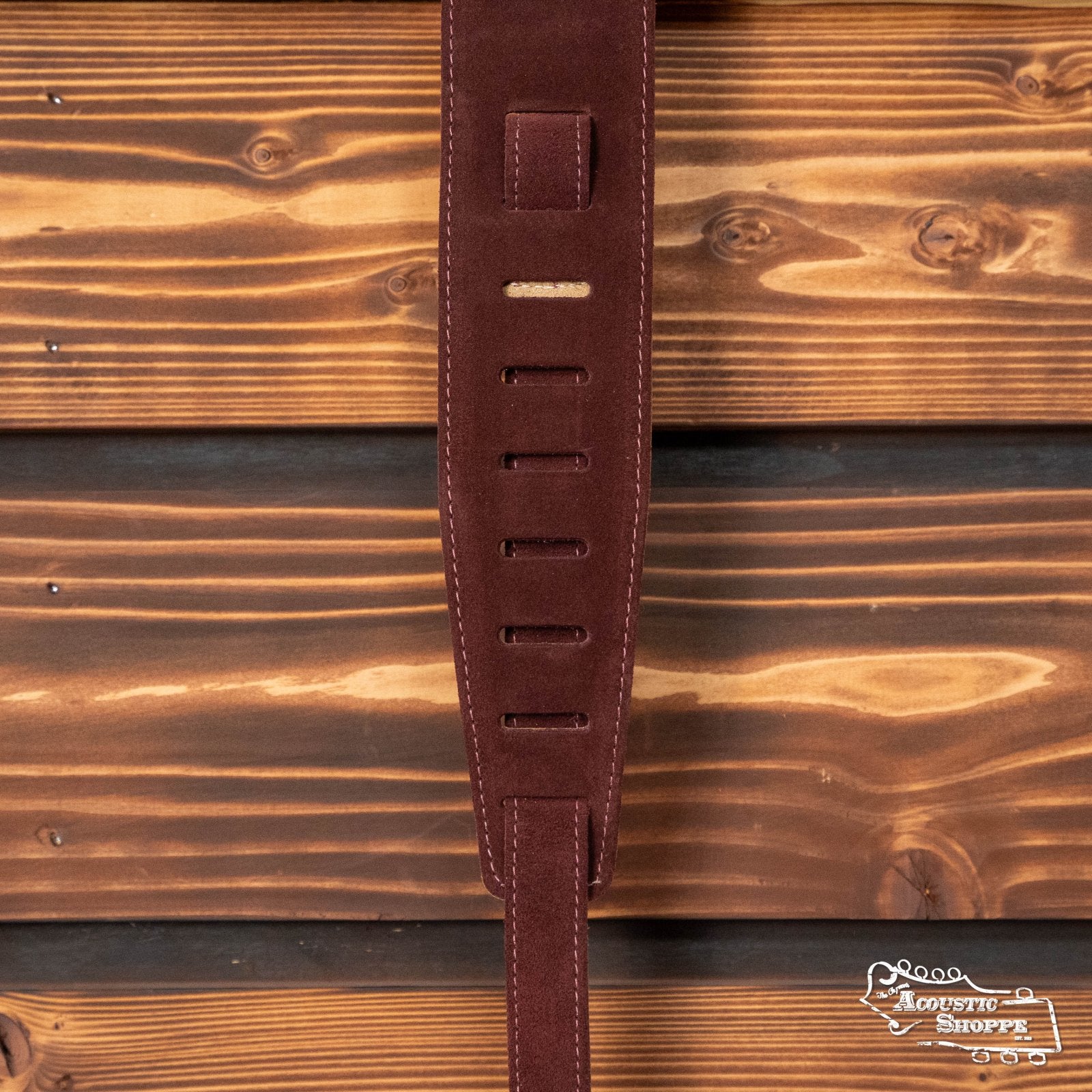 A D'Addario / Planet Waves Suede Guitar Strap in Burgundy (25SS03-DX) hangs vertically against a wooden background, with the Acoustik Shoppe logo visible in the lower right, highlighting this stylish accessory for musicians.