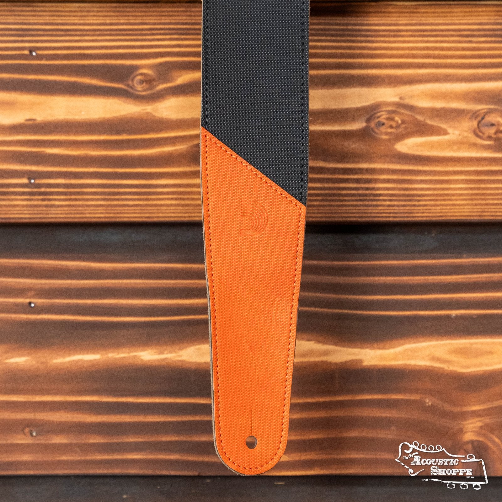 A close-up of a D'Addario 2.5" Leather Guitar Strap with orange ends (L25W1407) hangs against a wooden background, featuring The Acoustic Shoppe logo in the bottom right corner.