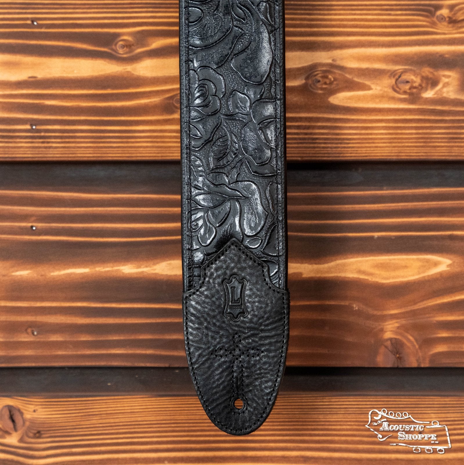 Levy's Leather 3" Embossed Leather Guitar Strap (M4WP-007) with floral detailing and garment leather backing hangs vertically against wood, featuring a padded, textured end and a small embossed logo near the strap button hole.