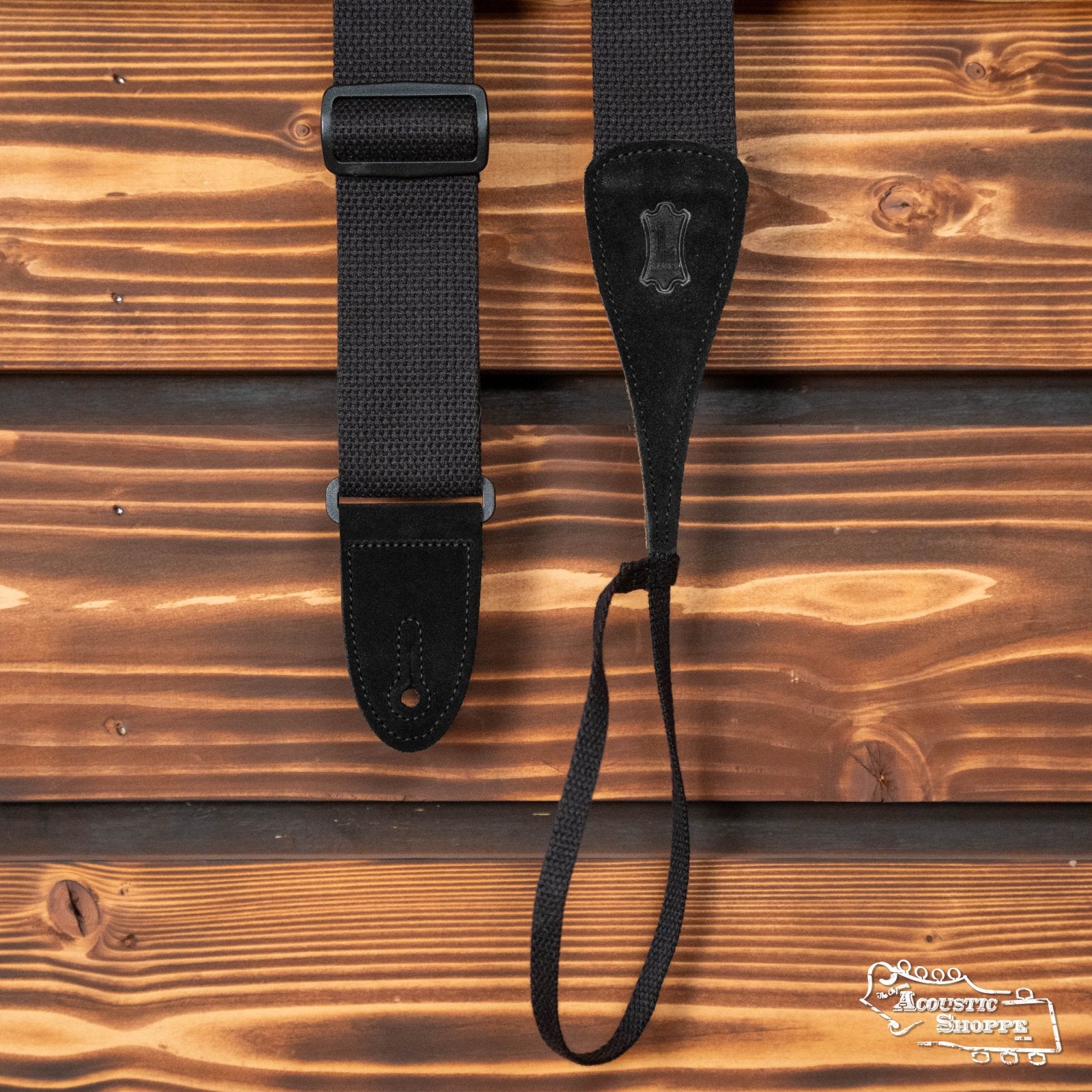 A Levys Leather Guitar/Dobro (MC8A-BLK) adjustable black cotton guitar strap with leather ends hangs vertically against a wooden background. The Acoustic Shoppe logo appears in the bottom right corner.