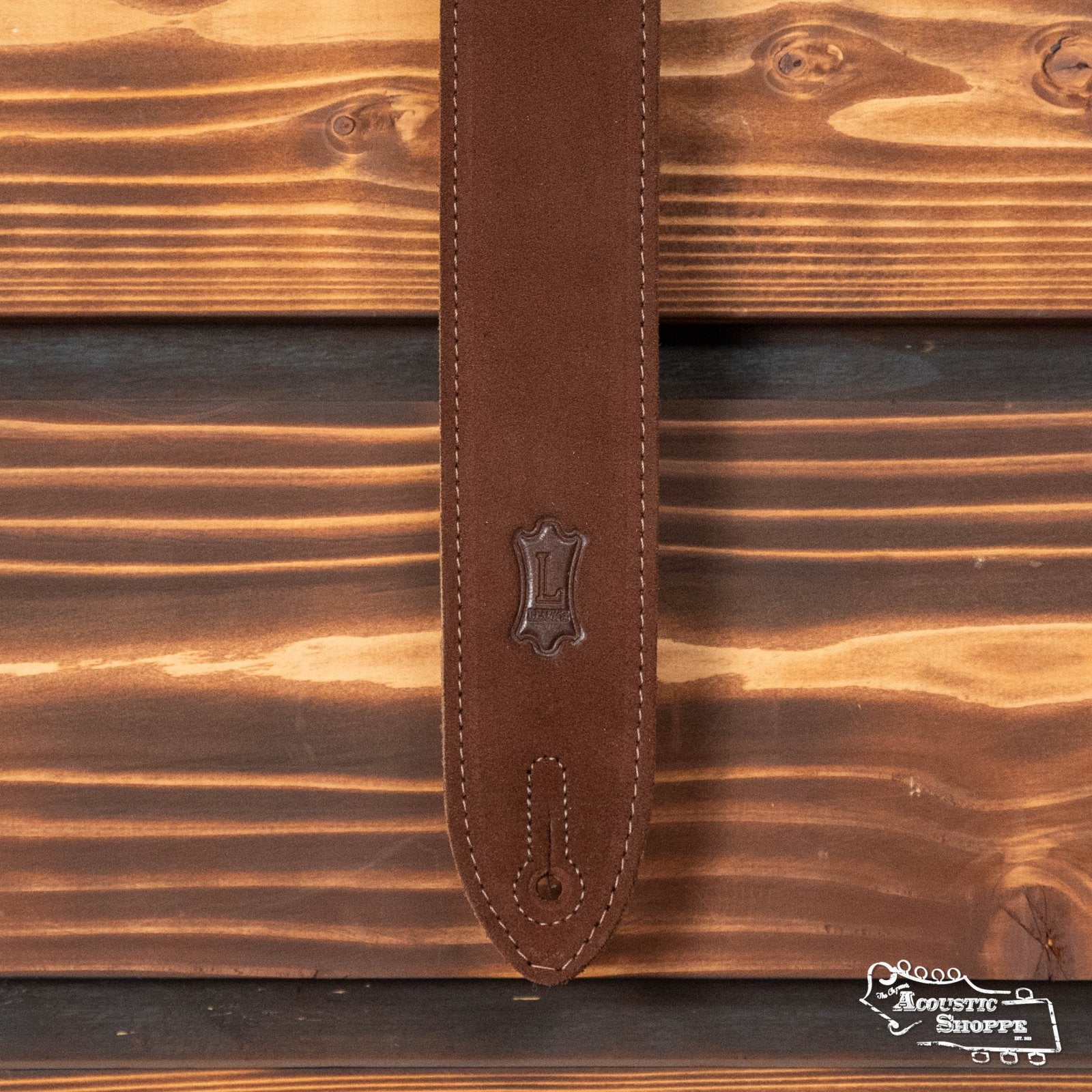 A Levys Leather 2 Suede Guitar Strap with suede backing in brown (MS12-BRN) is displayed against a wood-paneled background, featuring stitched edges and a leather emblem. The Acoustic Shoppe logo appears in the bottom right corner.