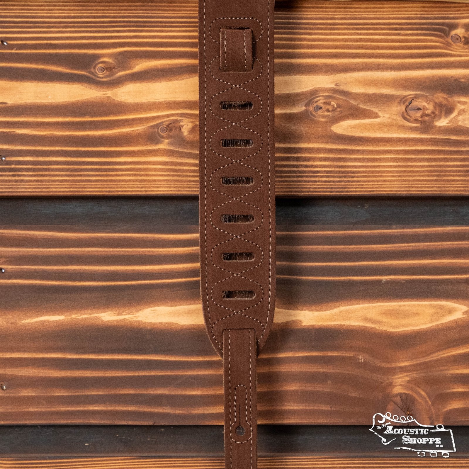 A Levys Leather 2 Suede Guitar Strap with suede backing (MS12-BRN) in brown hangs against a rustic wooden wall, featuring oval cutouts and decorative stitching. The Acoustic Shoppe logo appears in the bottom right corner.