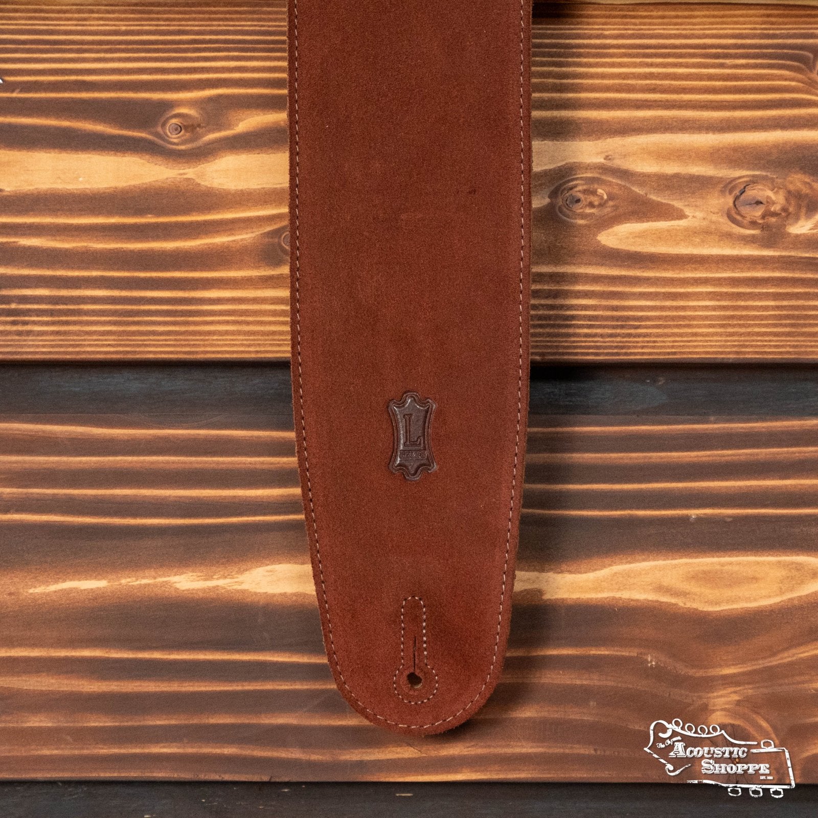 A Levys 3.5 Suede Leather Bass Strap With Suede Backing in Rust (MS4-RST) by Levys Leather with white stitching and an emblem rests on a wooden surface, featuring the Acoustic Shoppe logo in the lower right corner.