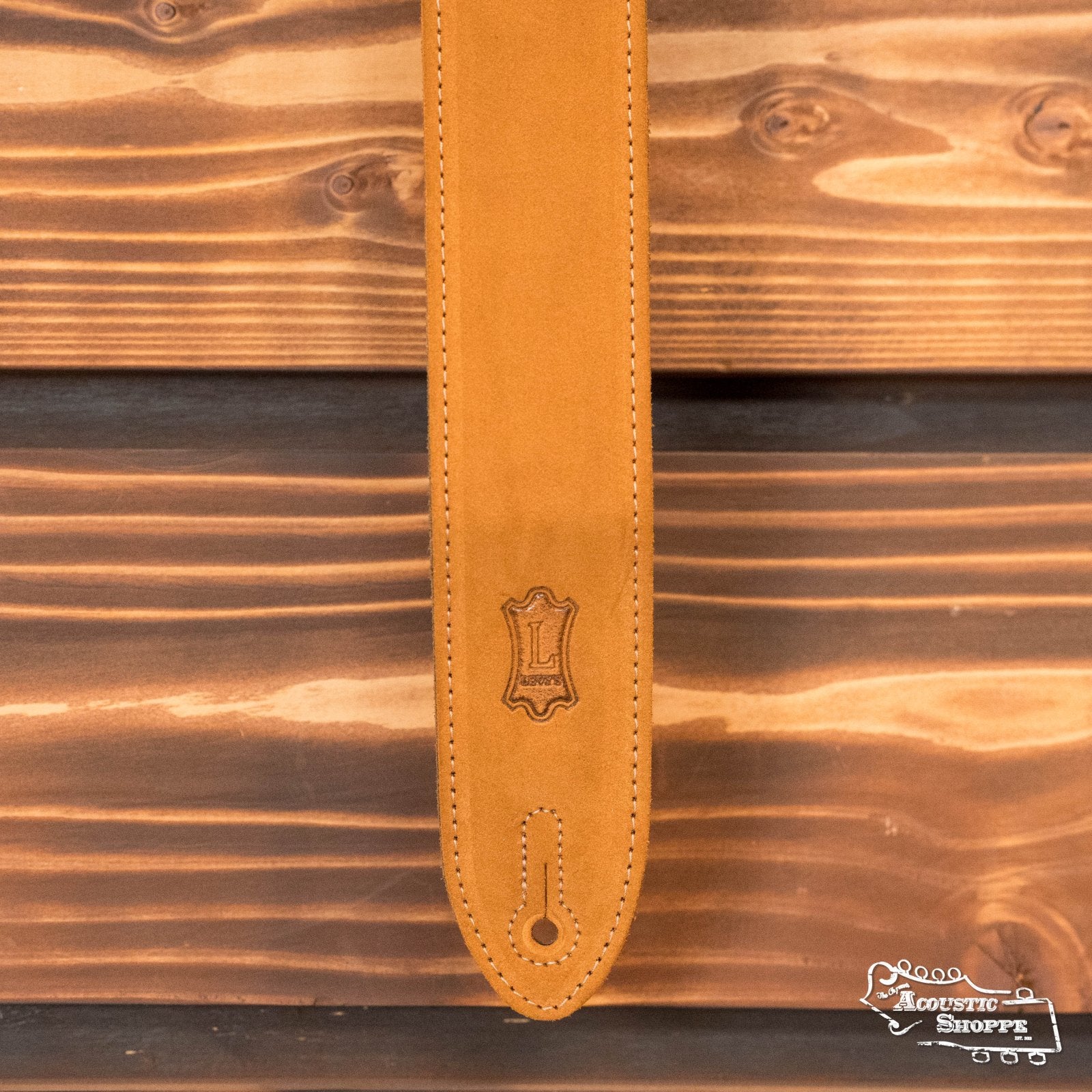 A Levys Leather 2 suede guitar strap in honey (MS12-HNY), featuring decorative stitching and an embossed L logo, is shown vertically on a wooden background, with The Acoustic Shoppe logo in the lower right corner.