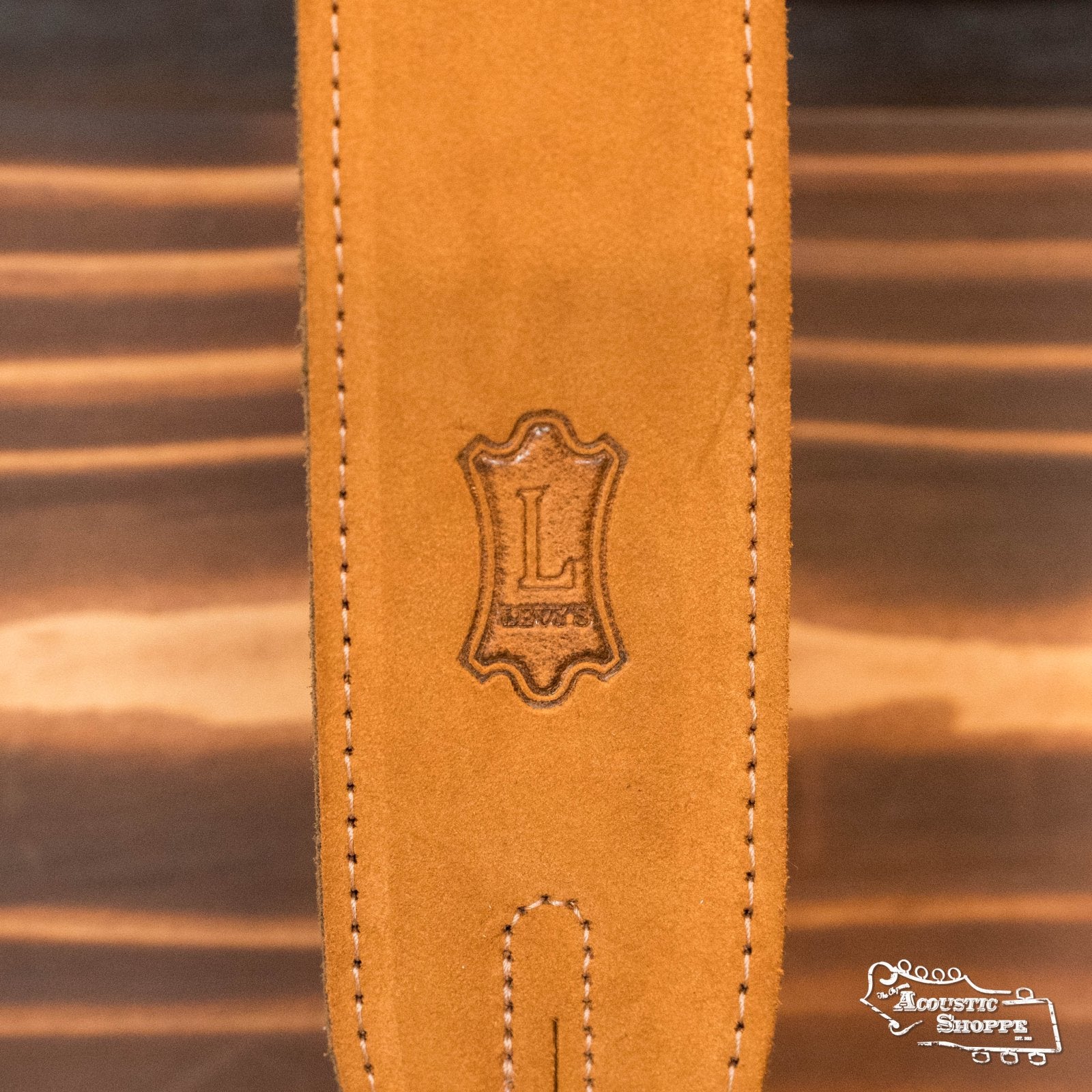 A close-up of the Levys Leather 2 Suede Guitar Strap in Honey (MS12-HNY) with an embossed L logo and 1973 in a decorative frame, set on a wooden surface featuring the Acoustic Shoppe logo.