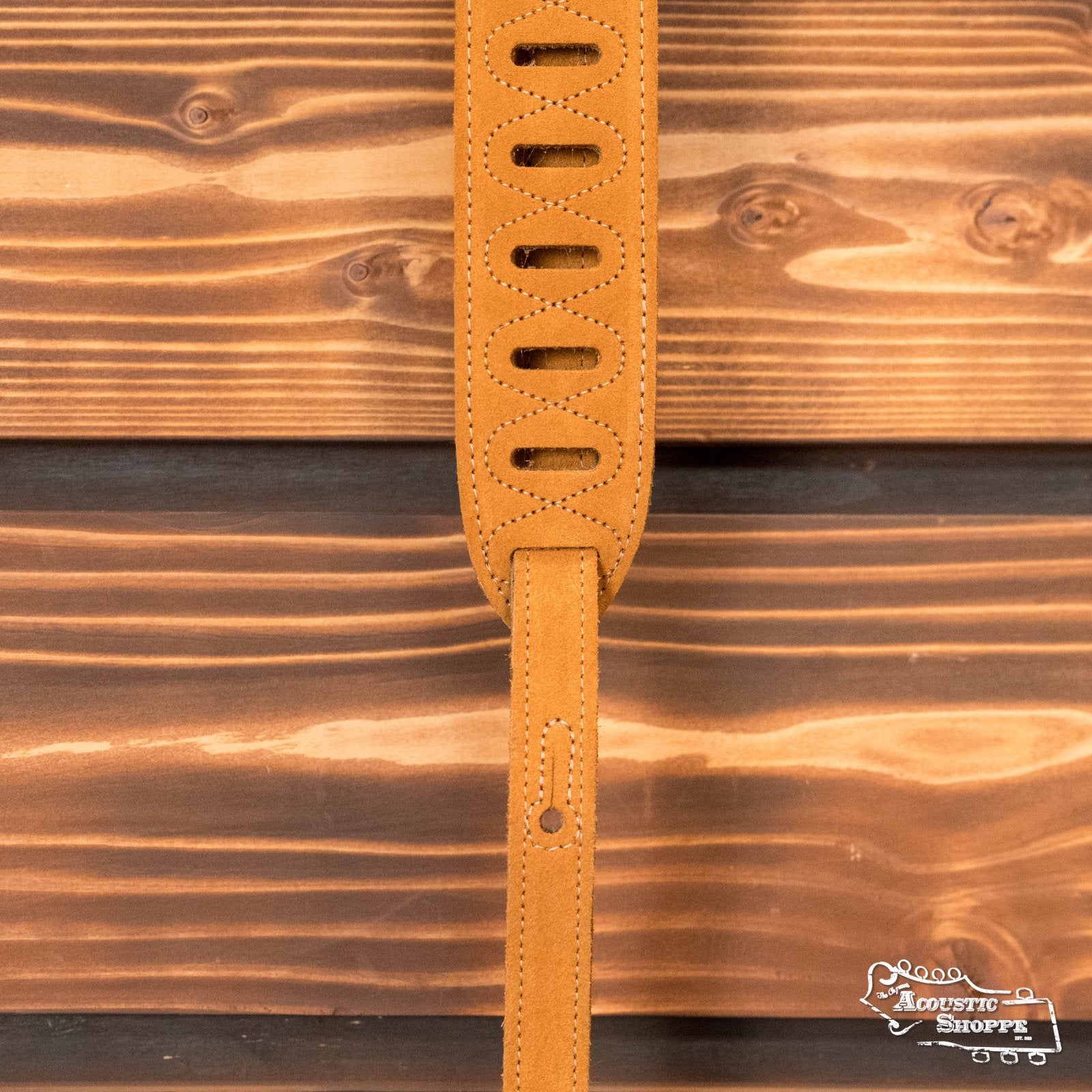 A Levys Leather 2 suede guitar strap in Honey (MS12-HNY) with oval cutouts is shown vertically against wood paneling. The Acoustic Shoppe logo appears in the lower right corner.