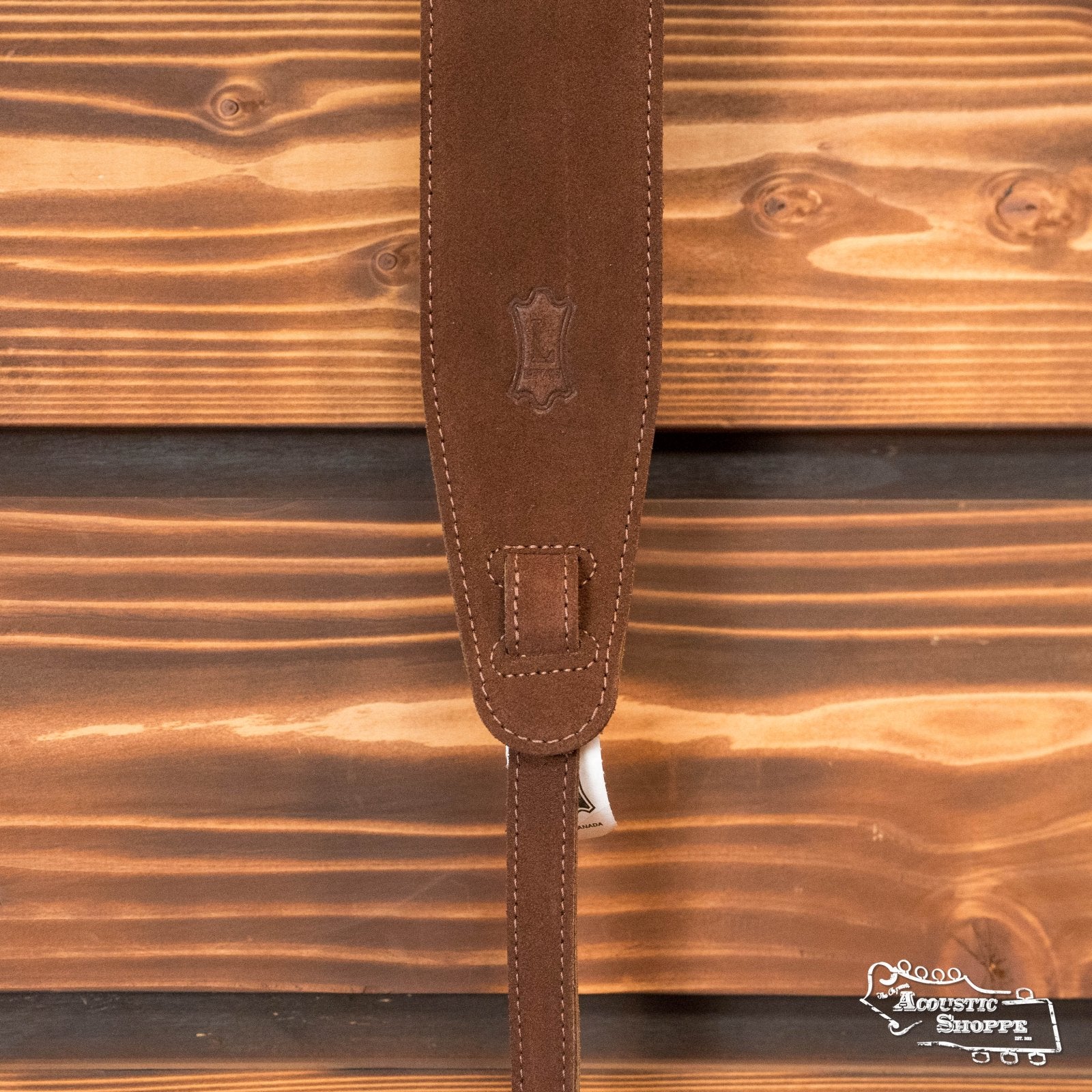 The Levys MS14-BRN Suede Leather Banjo Strap in brown by Levys Leather, featuring visible stitching and a leather emblem, hangs against a wooden background with The Acoustic Shoppe logo in the bottom right corner.