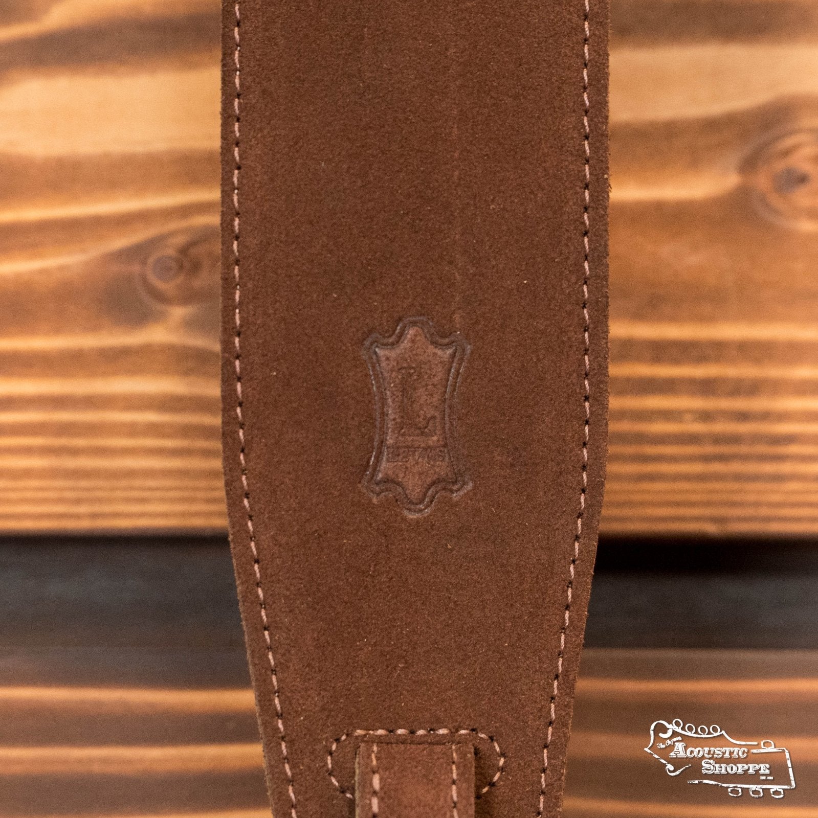Close-up of the Levys Leather MS14-BRN Suede Leather Banjo Strap in brown, featuring visible stitching and a central stamped emblem, displayed on a wooden surface. The Acoustic Shoppe logo appears in the bottom right corner.