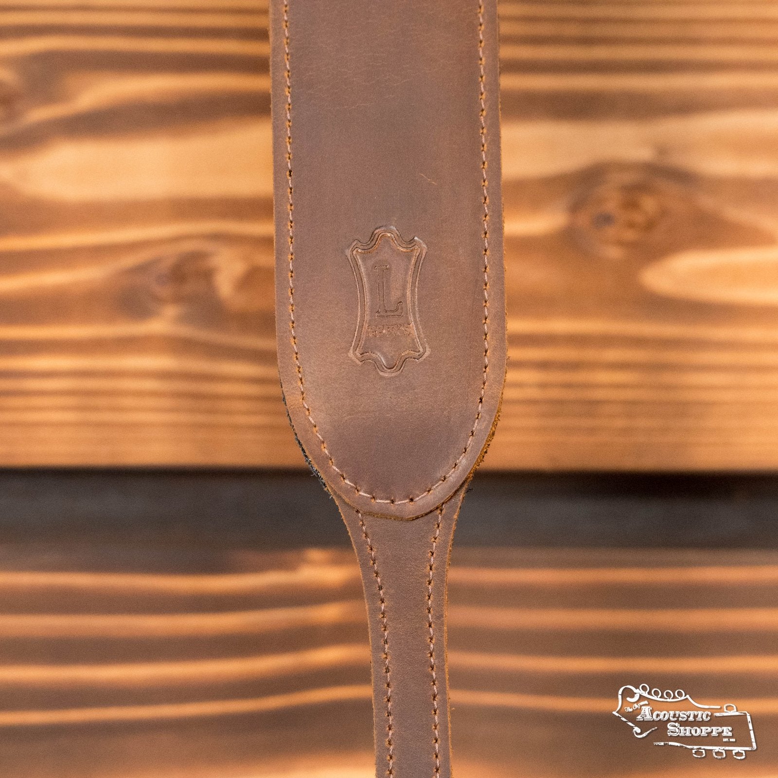 A close-up of the Levys Leather Banjo Master Series PMB32GF-BRN Padded Butter Leather Banjo Strap in brown, featuring decorative stitching and an embossed emblem on a wooden background with The Acoustic Shoppe logo in the lower right corner.