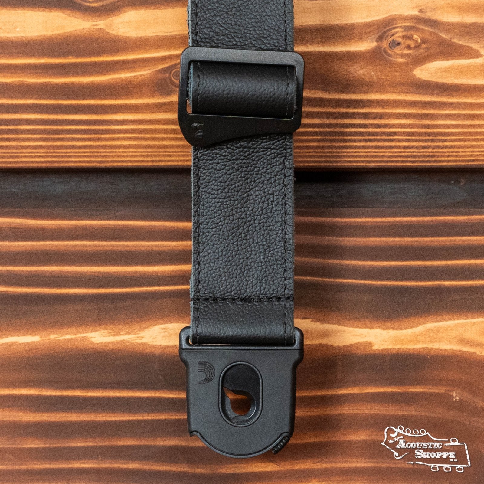 The DAddario / Planet Waves 50PLL00 Planet Lock Leather Guitar Strap in black is displayed on a wooden surface, featuring The Acoustic Shoppe logo in the bottom right corner.