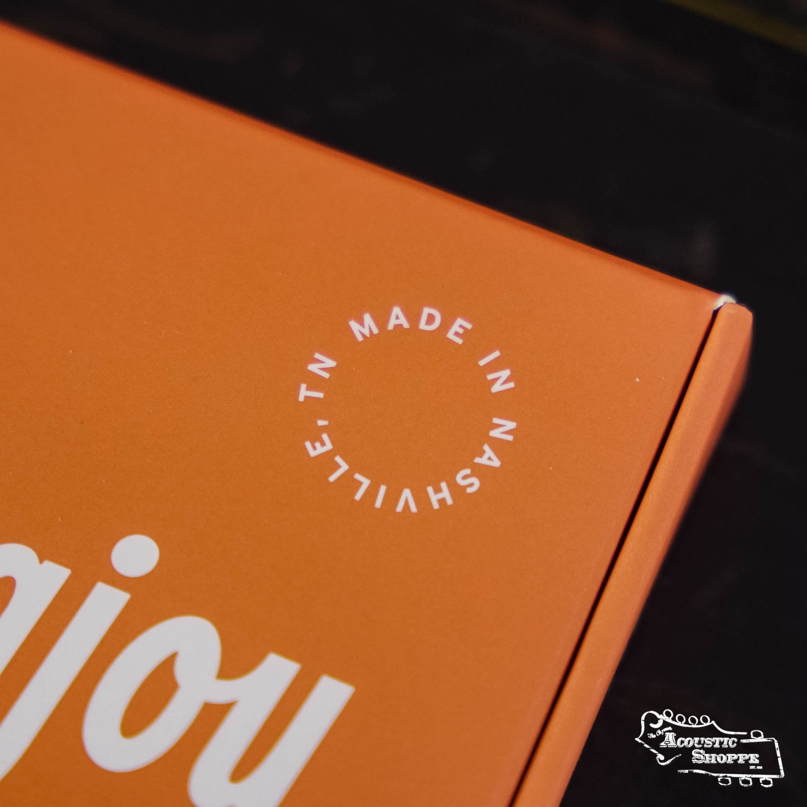 An orange box with white text reading “MADE IN NASHVILLE, TN” in a circle, part of "enjoy," a small white Stringjoy Acoustic Shoppe logo, and reference to Stringjoy Foxwoods Medium Gauge (13-56) coated phosphor bronze acoustic guitar strings appear.