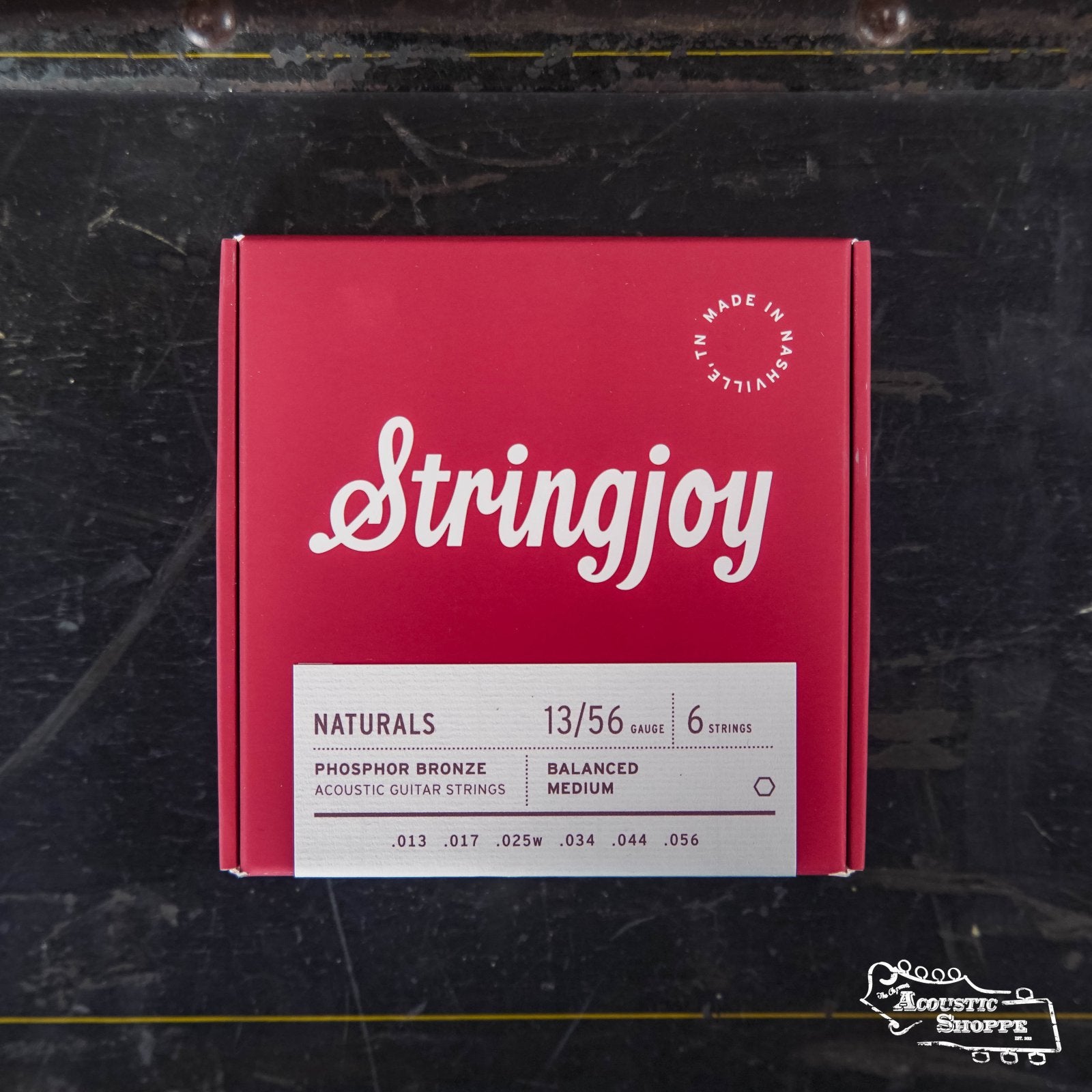 A red Stringjoy Naturals Medium Gauge (13-56) Phosphor Bronze Acoustic Guitar Strings box rests on a dark, worn surface, with the Acoustic Shoppe logo visible in the lower right corner.