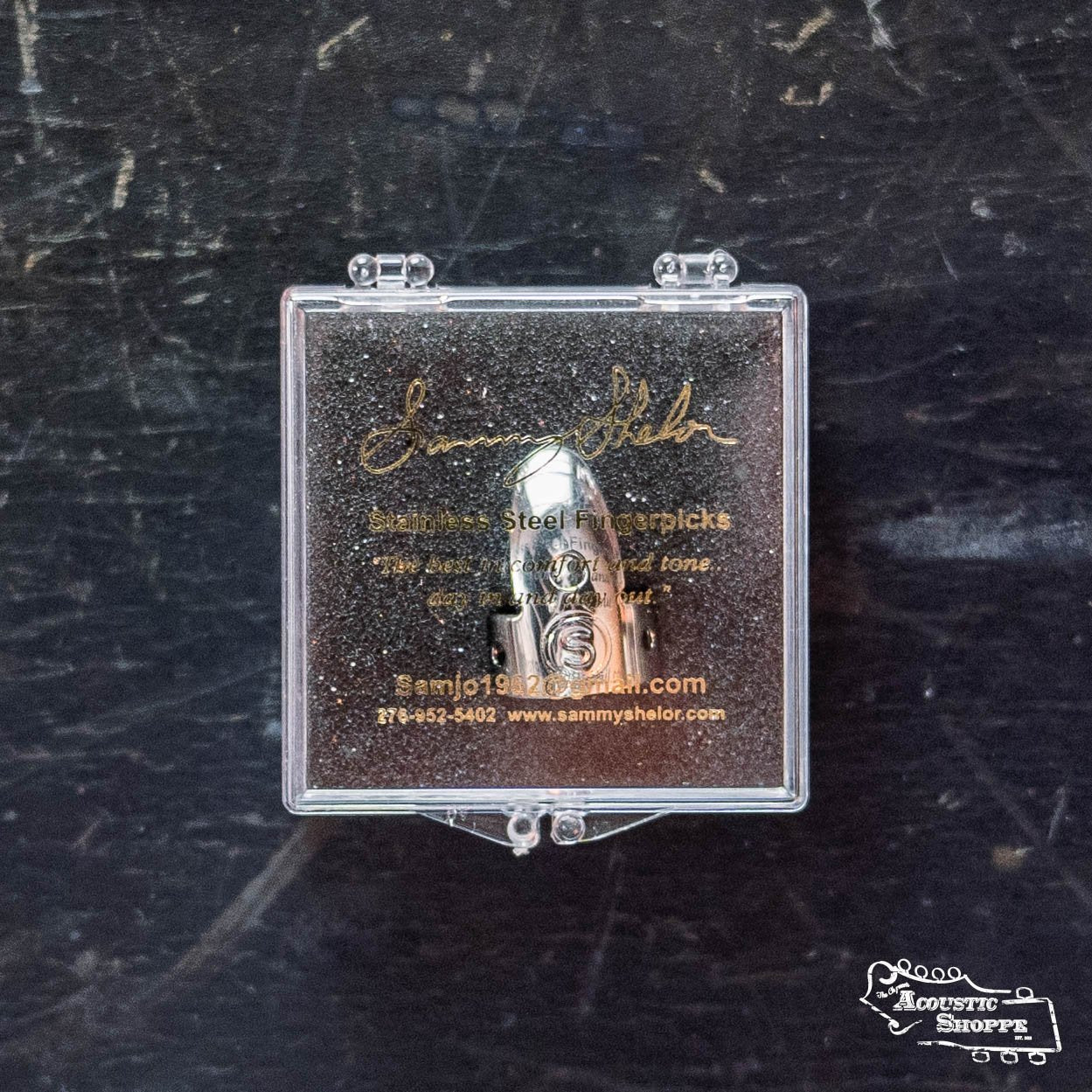 The Sammy Shelor Stainless Steel Fingerpicks, Wide Band are showcased in a clear case with gold lettering on a dark surface, featuring The Acoustic Shoppe logo—a top pick for fingerstyle guitarists.