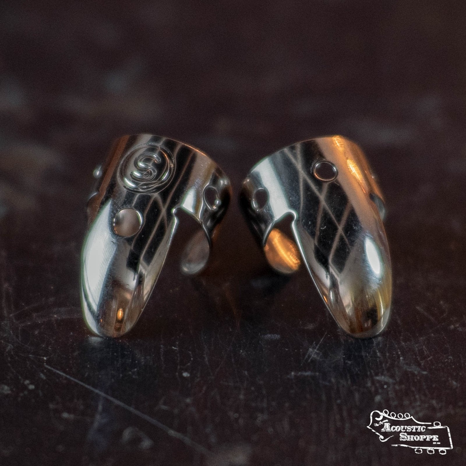 Two Sammy Shelor Stainless Steel Fingerpicks (Wide Band) lie side by side on a reflective surface. Each pick features small circular cutouts, with one engraved swirl. The Acoustic Shoppe logo appears in the bottom right corner.