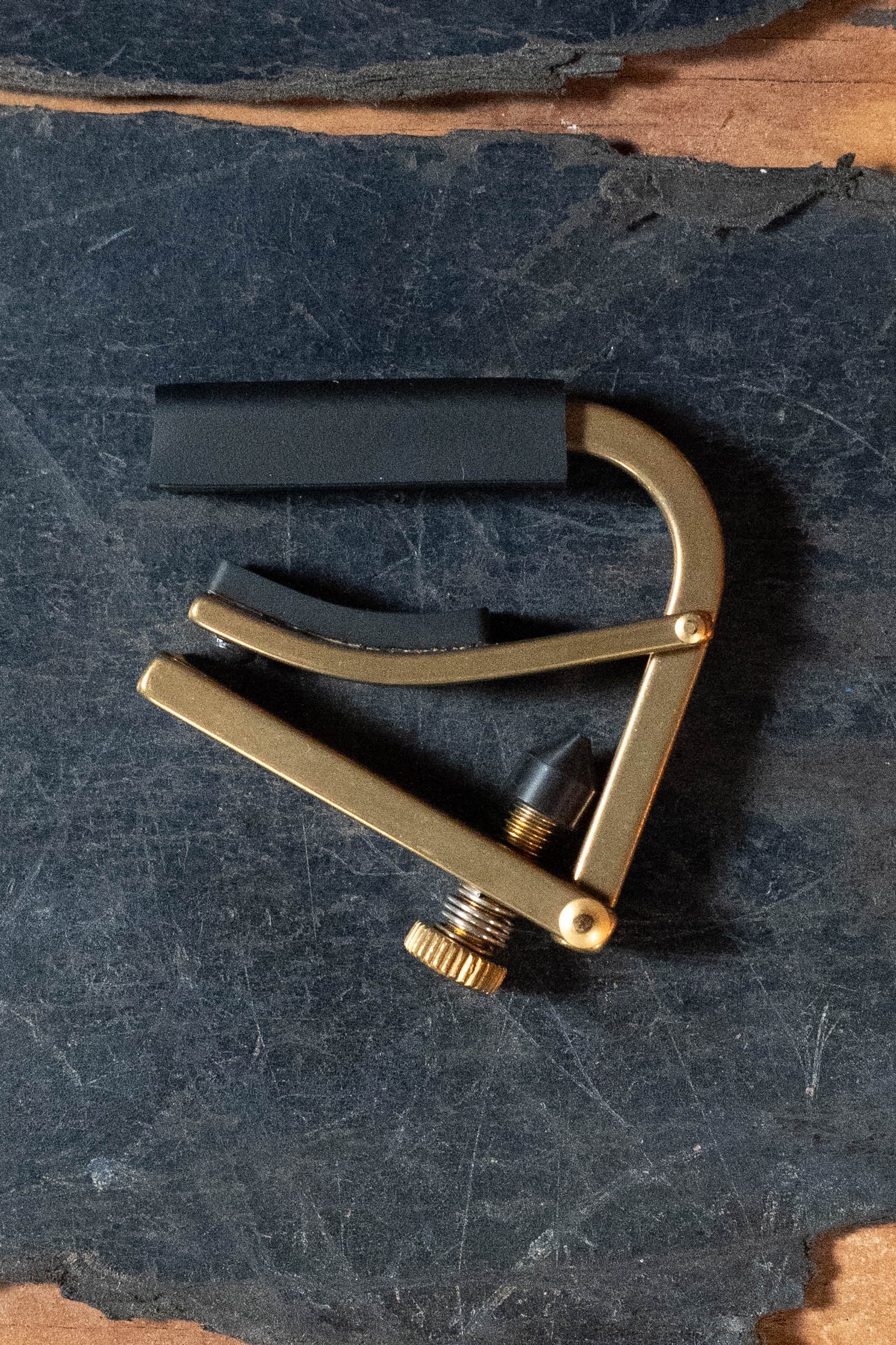 A Shubb C5b Classic Brass Banjo Capo by Shubb, featuring a gold finish and black rubber clamp for flat fretboards, rests on a dark, textured surface with rough brown edges.