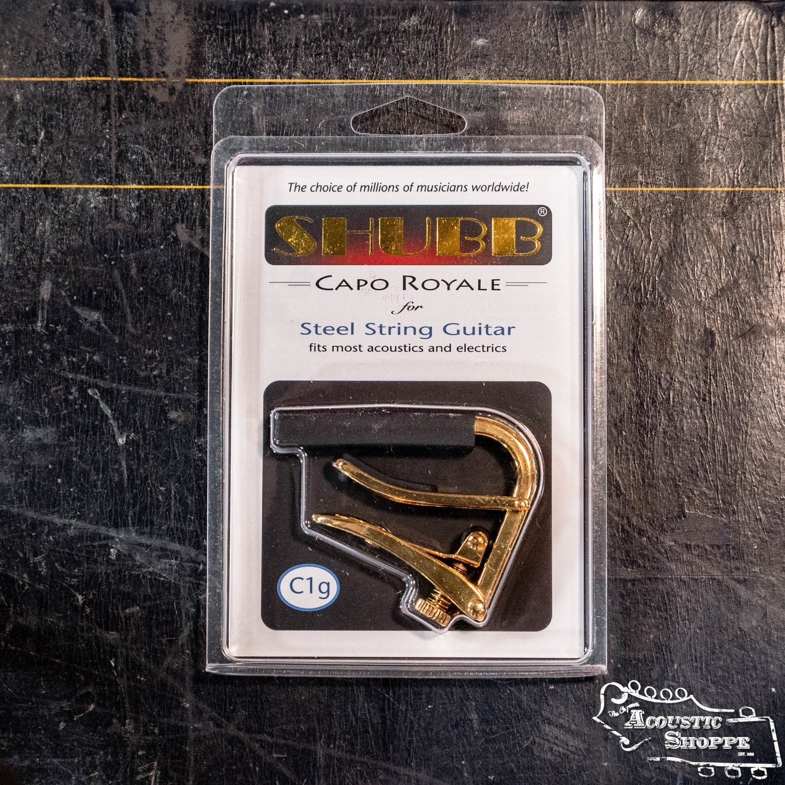 The Shubb C1g Royale Gold Guitar Capo by Shubb is shown in clear plastic packaging on a dark surface, with the Acoustic Shoppe logo visible in the bottom right corner.