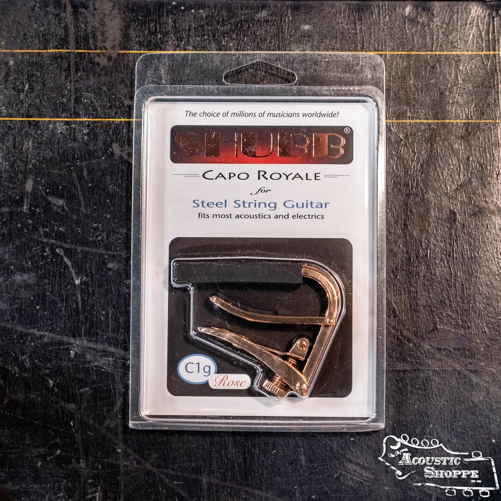 A Shubb C1rg Royale Rose Gold Guitar Capo by Shubb, in original packaging labeled “fits most acoustics and electrics,” rests on a dark, textured surface with Acoustic Shoppe branding in the corner.