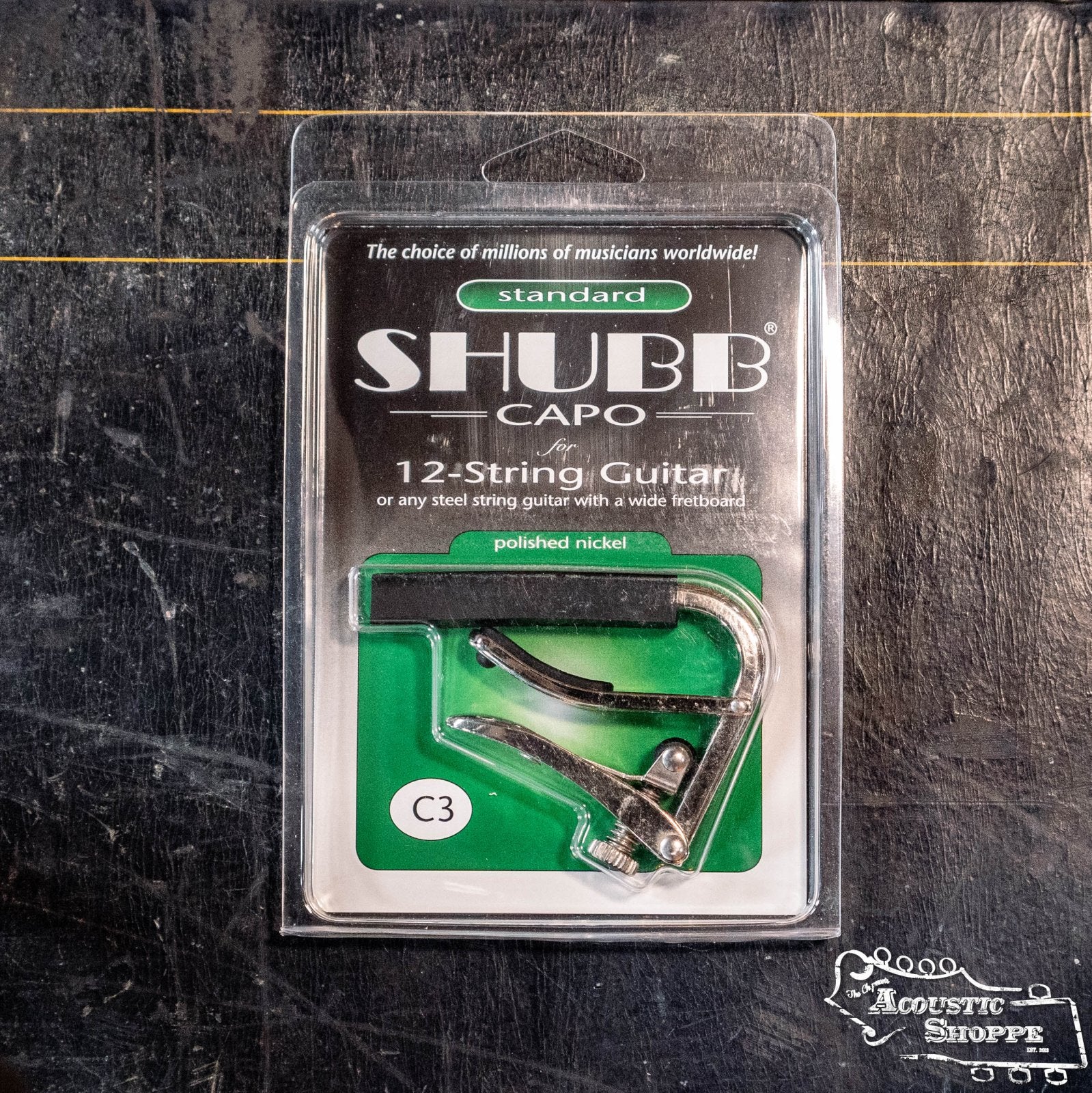 A packaged Shubb C3 Nickel 12-String Guitar Capo from Shubb is shown on a dark textured surface with the Acoustic Shoppe logo in the bottom right corner. Ideal for steel string guitars.