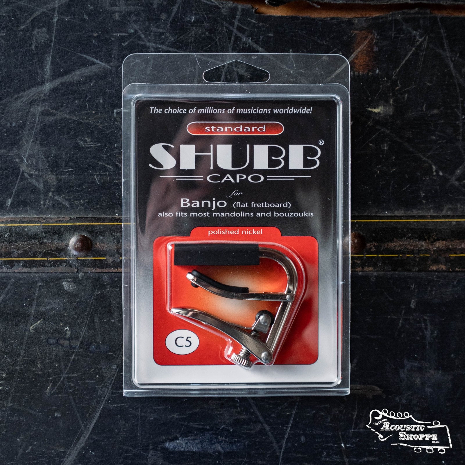 A Shubb Classic Banjo Capo (C5) by Shubb with a polished nickel finish for banjo, mandolin, and bouzouki is shown in its red, black, and white packaging on a dark textured surface, highlighting the over-center locking mechanism.