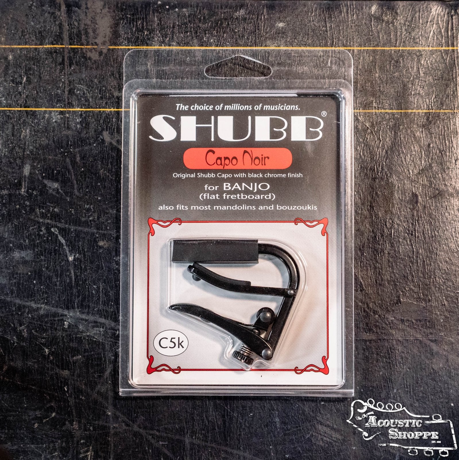 A Shubb Capo Noir Banjo (C5K) by Shubb, with a black chrome finish and over-center locking mechanism, is shown in its packaging on a dark textured surface. The Acoustic Shoppe logo appears at the bottom right.