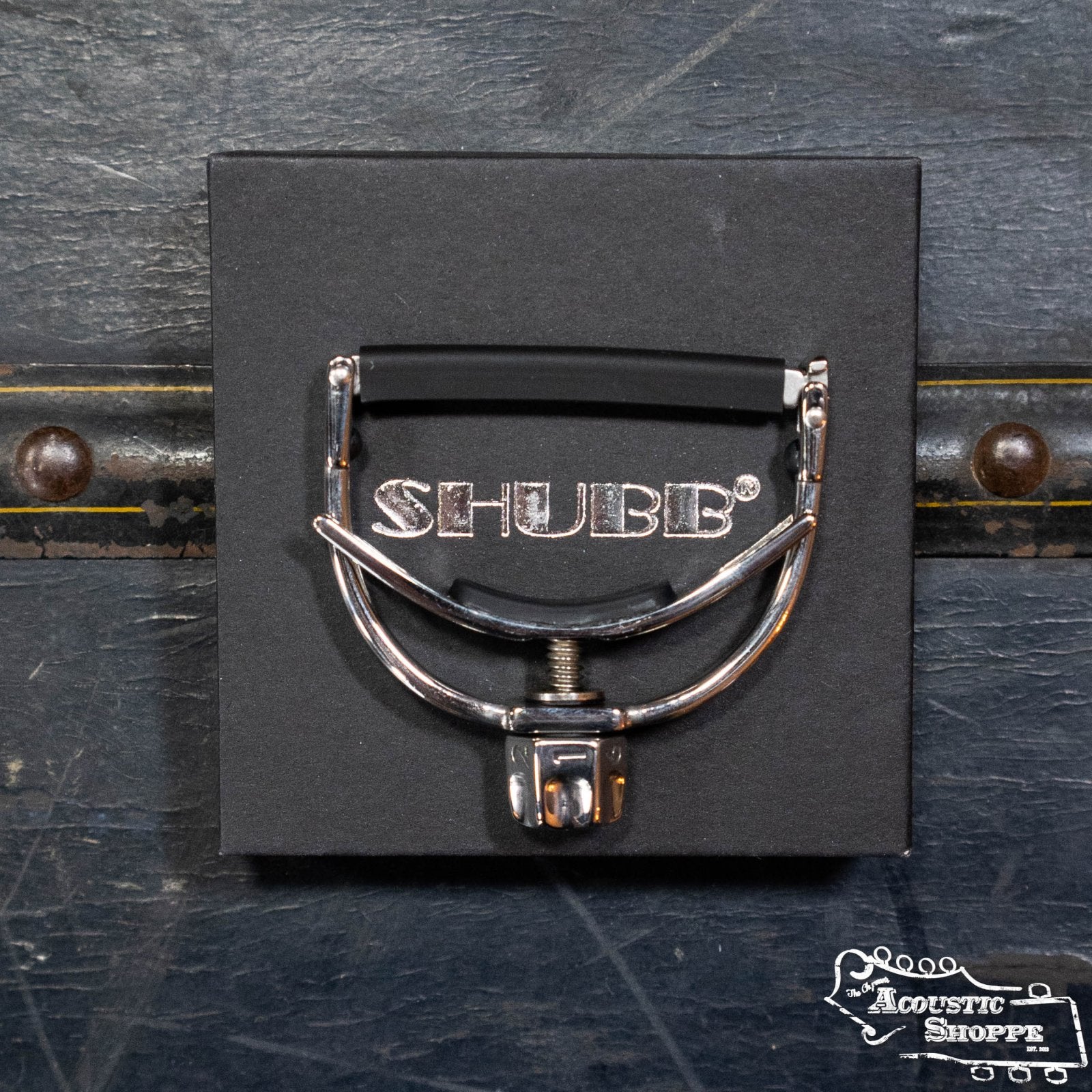 A Shubb F1 FineTune Screw-Operated Guitar Capo for steel string guitars is shown on black Shubb packaging atop a dark textured surface, with The Acoustic Shoppe logo in the lower right corner.