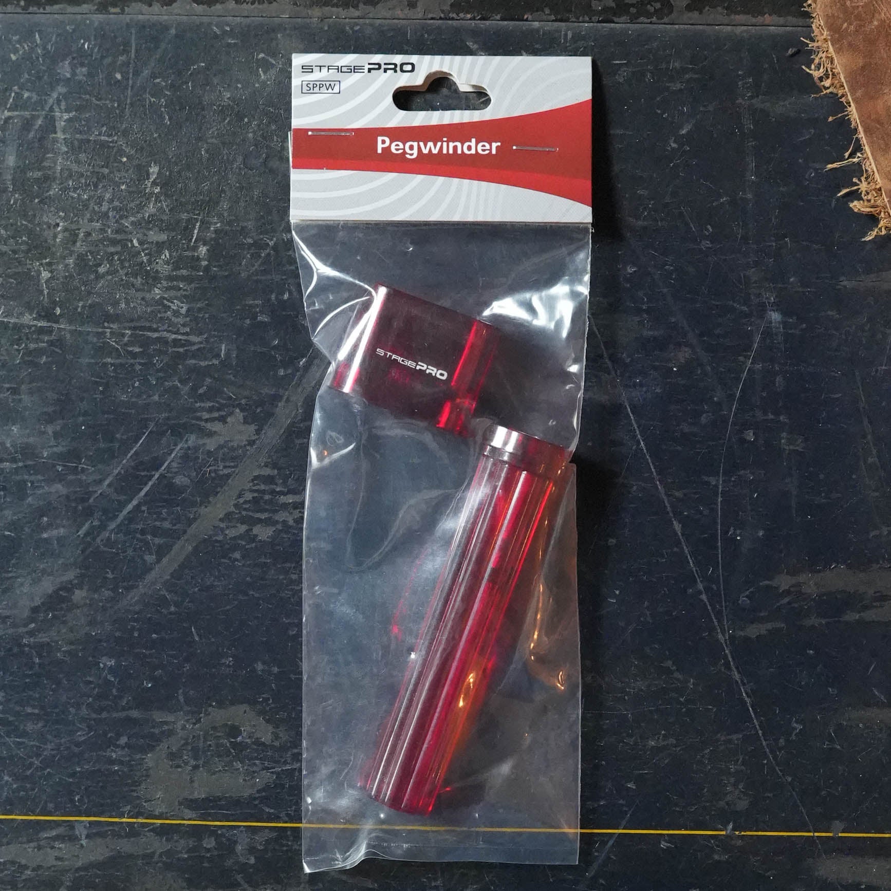 A StagePro pegwinder by The Acoustic Shoppe, featuring a red handle and black top for tuning stringed instruments, is shown packaged on a dark textured surface.
