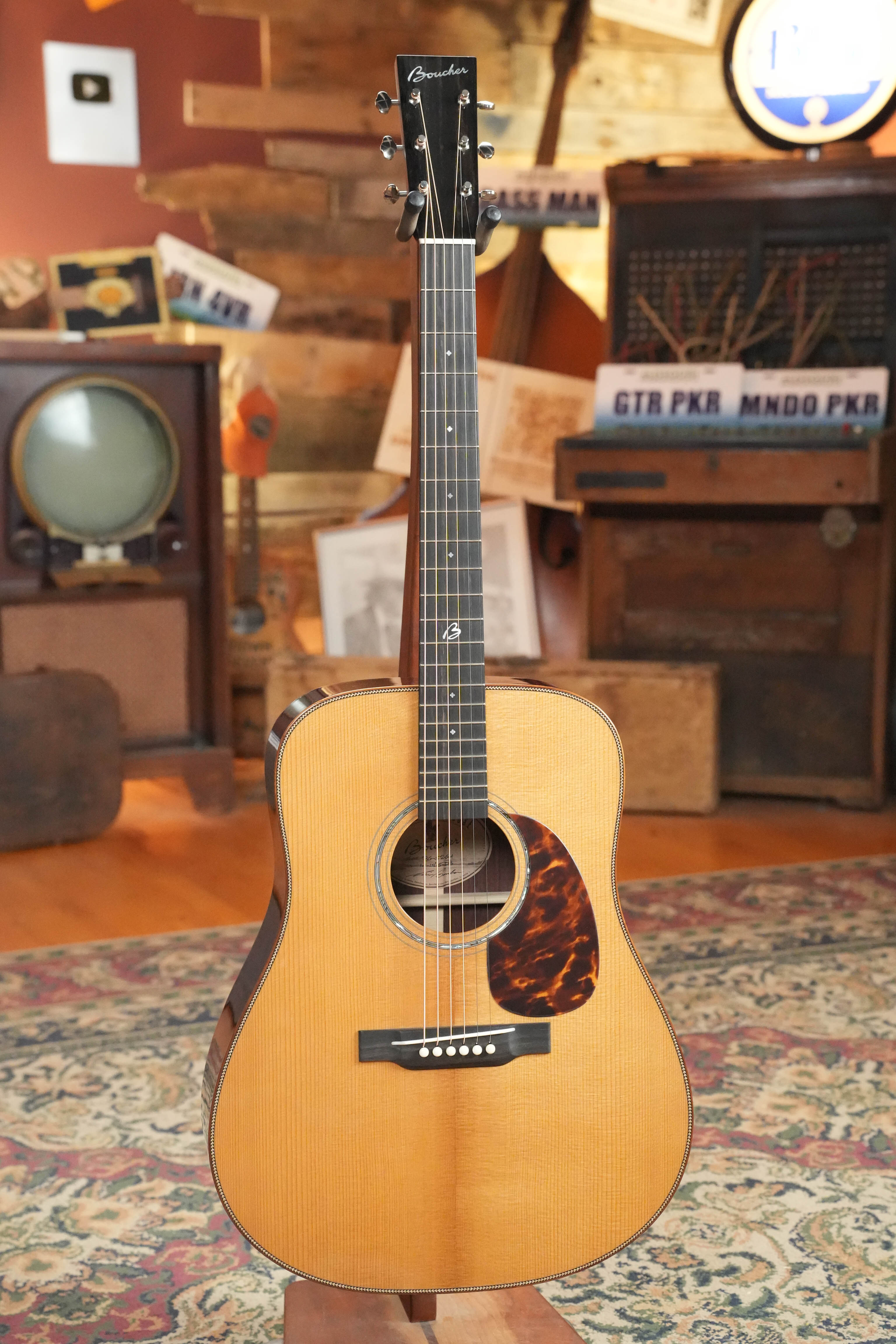 A *Stage Ready* Boucher BG-52-GM Torrefied Adirondack/Rosewood Bluegrass Goose Gold Master Pack Dreadnought Acoustic Guitar #1415 by Boucher Guitars stands on a patterned rug amid vintage electronics, signs, and eclectic decor—minor cosmetic flaws noted.