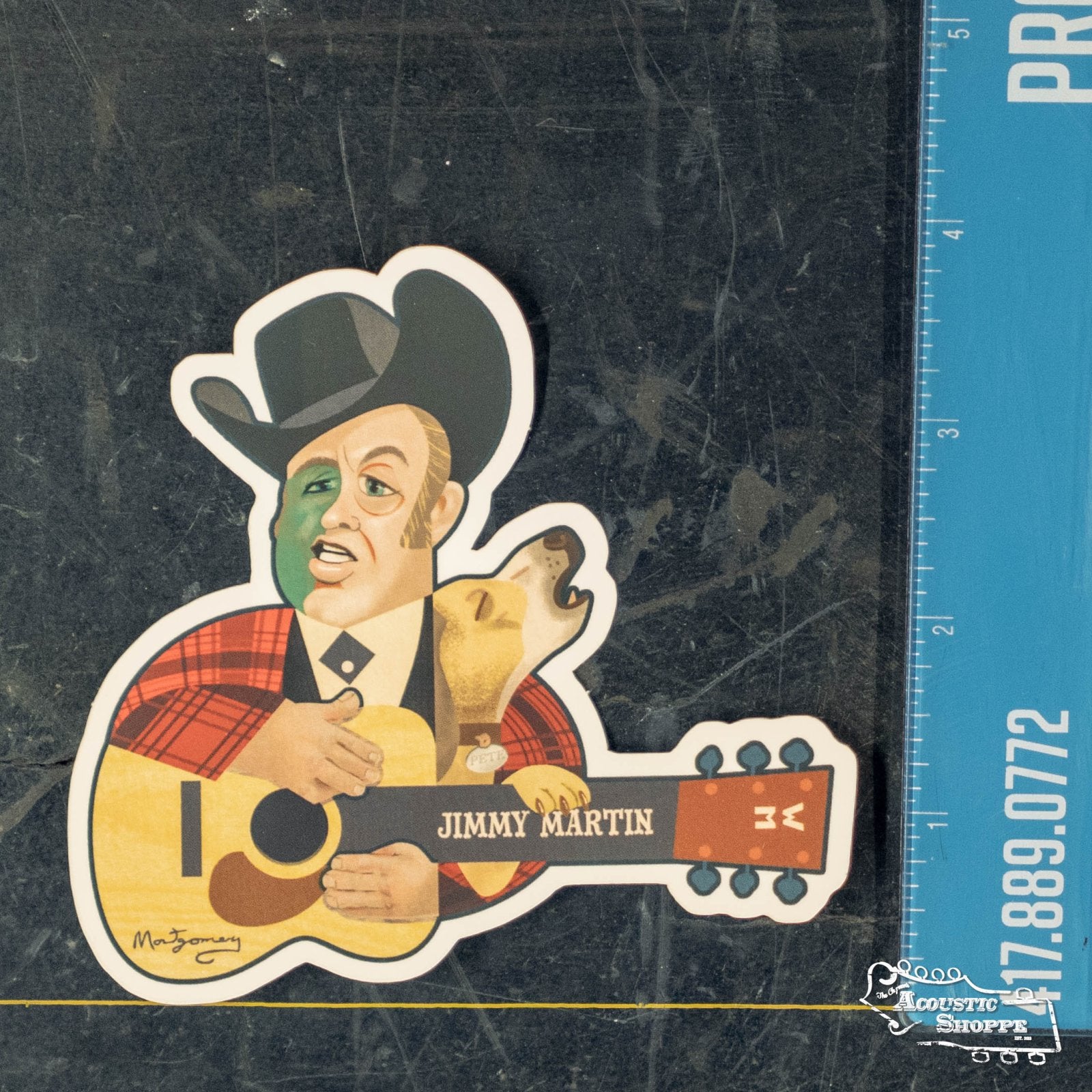 The Acoustic Shoppe’s Jimmy Martin Sticker by Mark A Montgomery features a man in a cowboy hat and red plaid jacket playing guitar with a dog—ideal for music fans. Shown next to a blue ruler for size reference.
