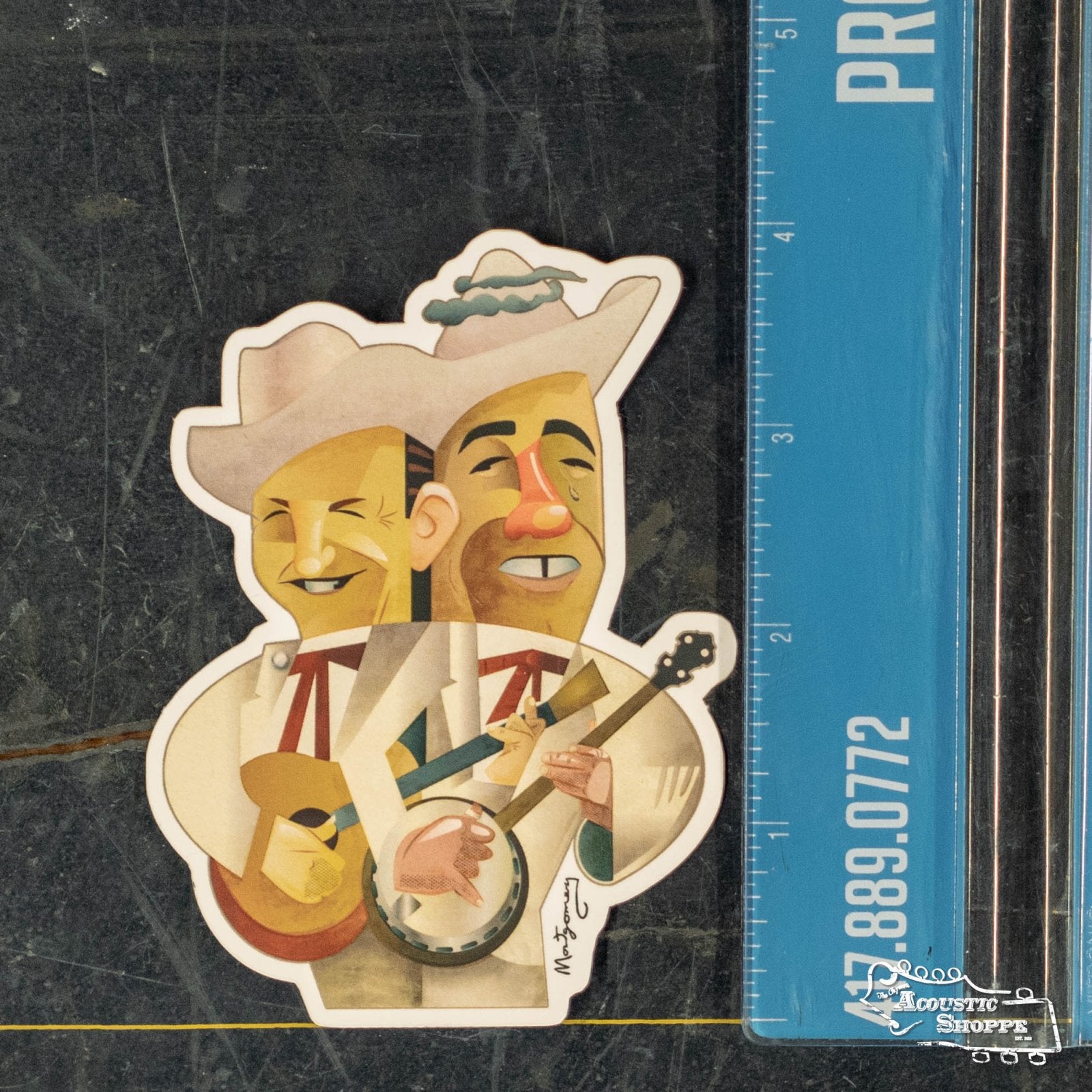 The Flatt & Scruggs Fine Art Sticker by Mark A Montgomery, showing two smiling cartoon musicians in hats with acoustic instruments, is placed beside a ruler on a dark, marbled surface for size reference. Brand: Mark A Montgomery.