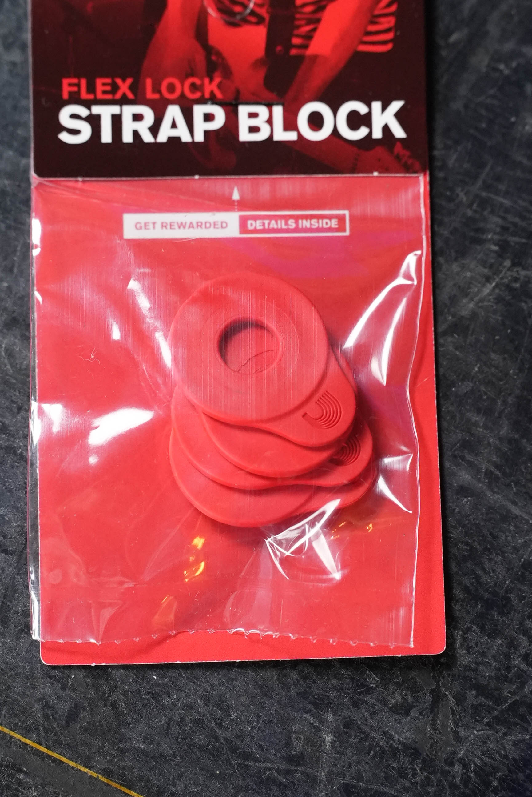 A D'Addario Flex Lock Strap Block 4-Pk - Red contains four red circular rubber blocks for securing guitar straps to end pins. The D'Addario-branded package shows the blocks through clear plastic, displayed on a dark surface.